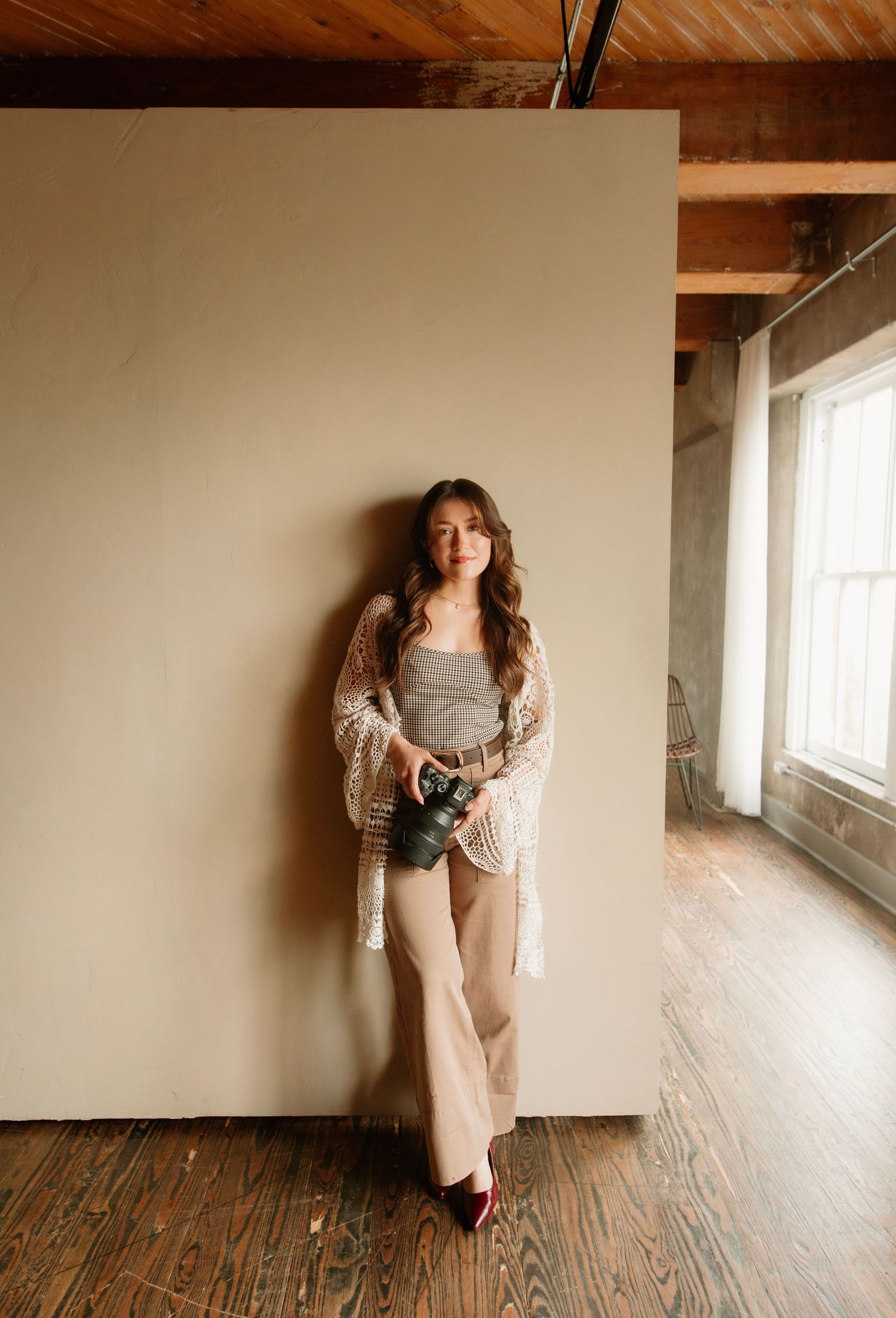 Woman in crochet cardigan and wide-leg pants holds camera, leaning against cream wall in rustic studio space.