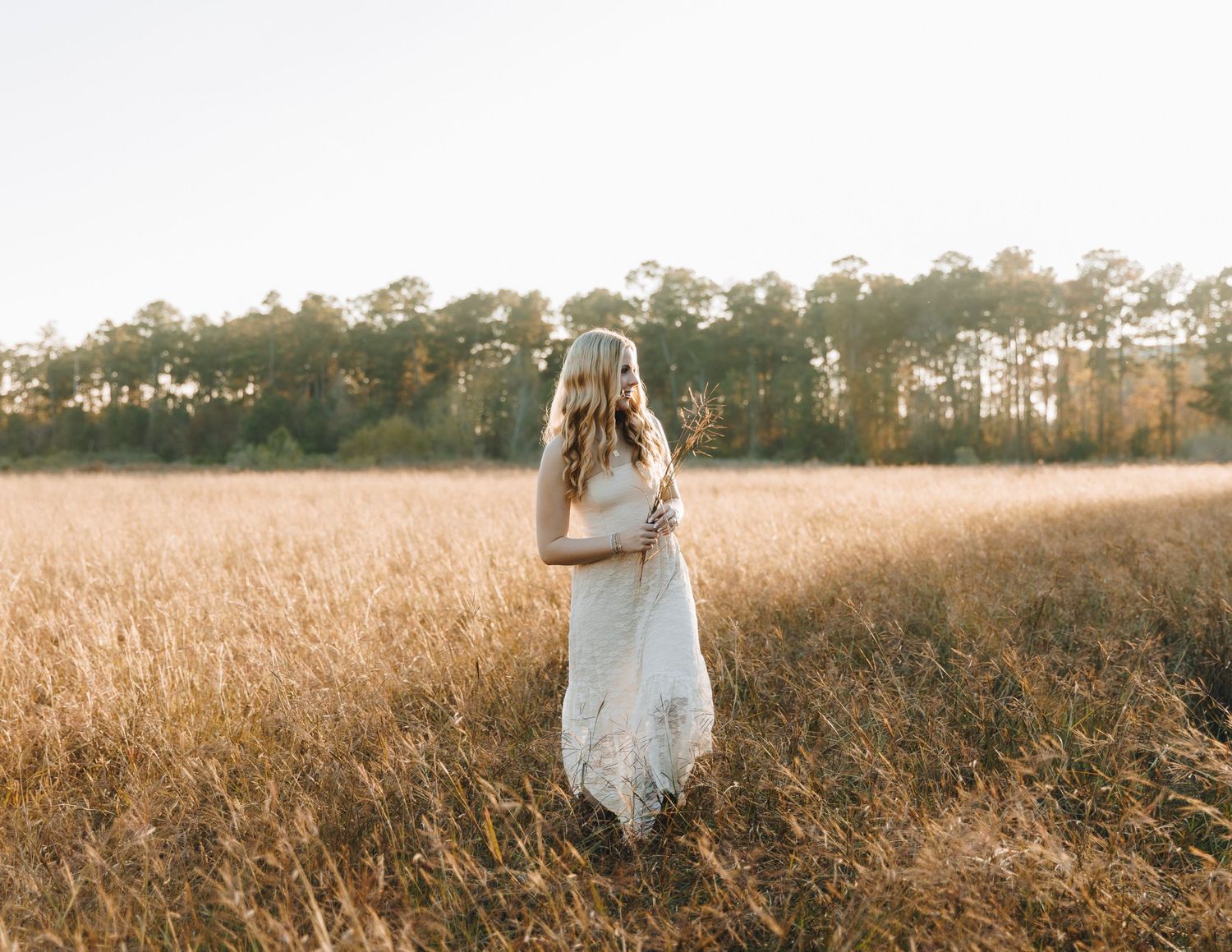 Blonde woman in white lace dress standing alone in golden field with trees in background.