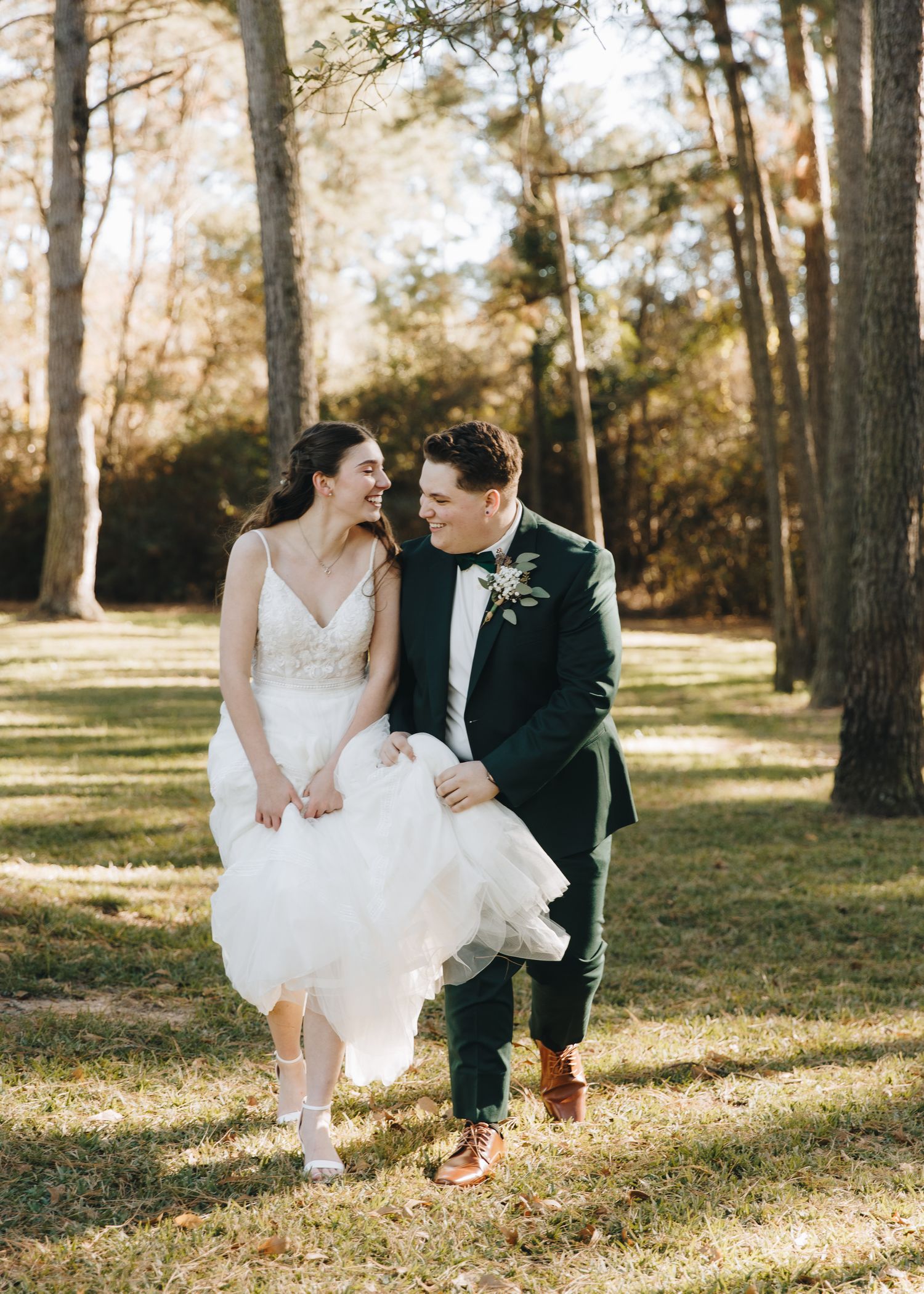Laughing bride and groom walk hand in hand through sunlit pine forest during joyful outdoor wedding portrait session.