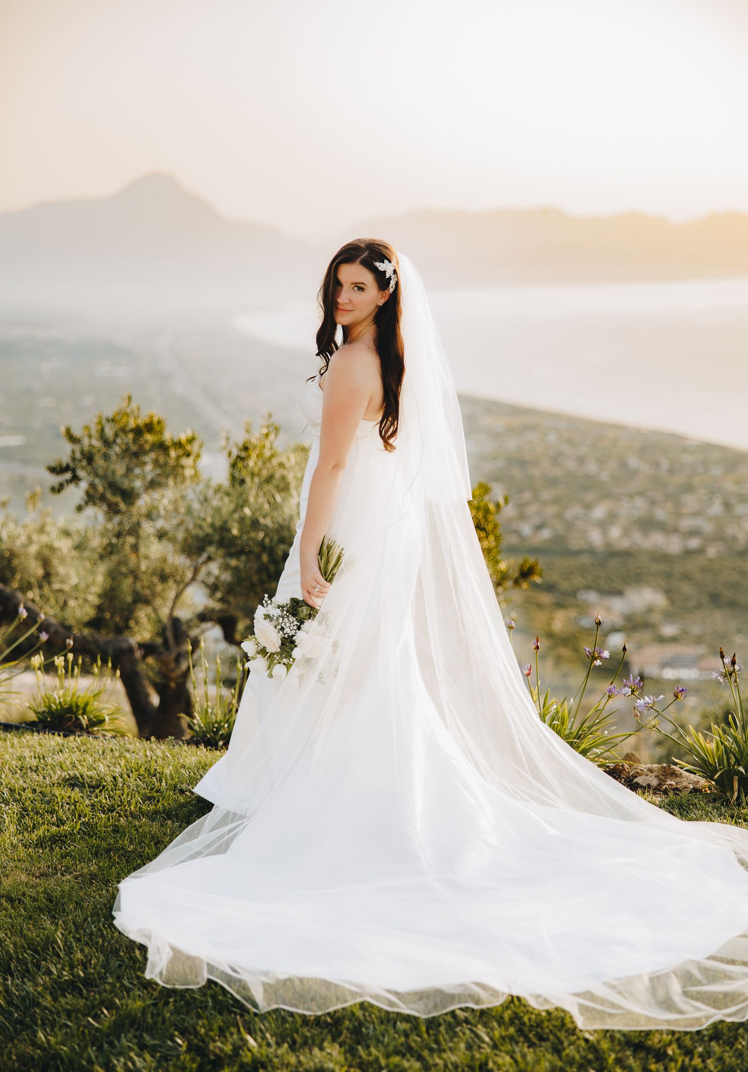 Bride in flowing white gown and veil poses with bouquet against stunning mountain and sea backdrop.