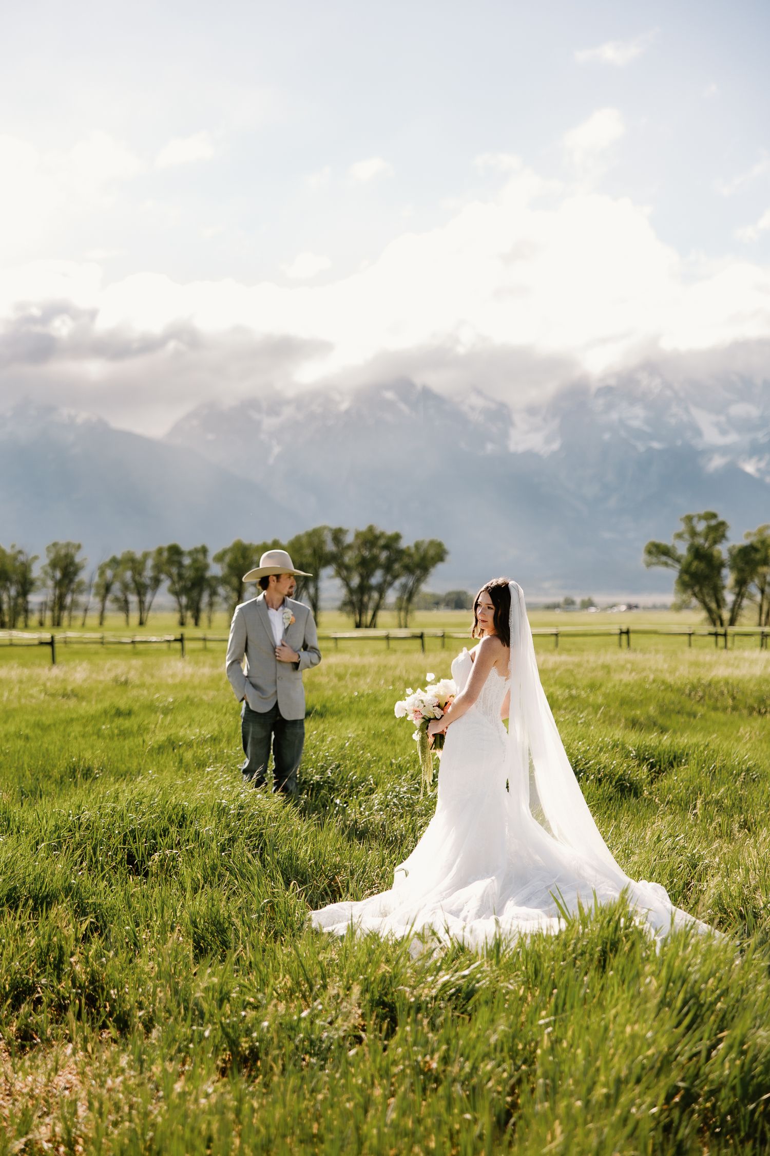 Bride in white gown with veil and groom in hat stand in green meadow with Grand Teton mountains backdrop.