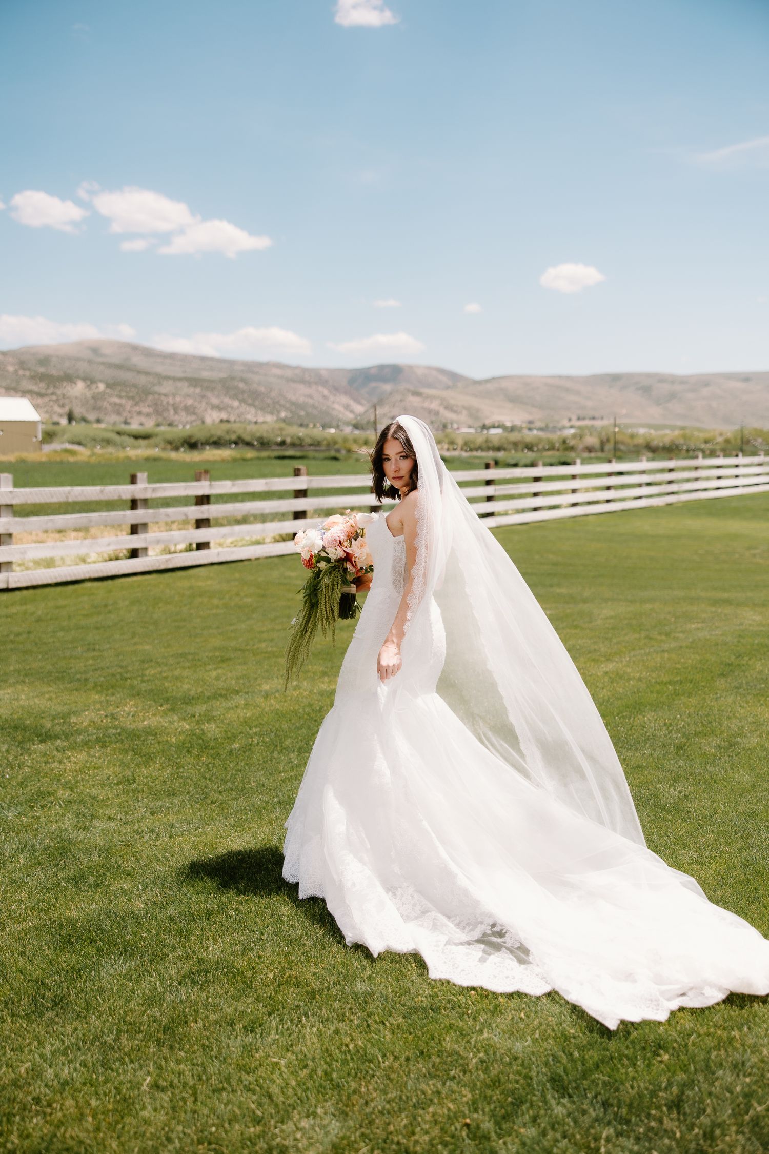 Bride in white gown and veil holding bouquet on green lawn with white fence and mountain backdrop.