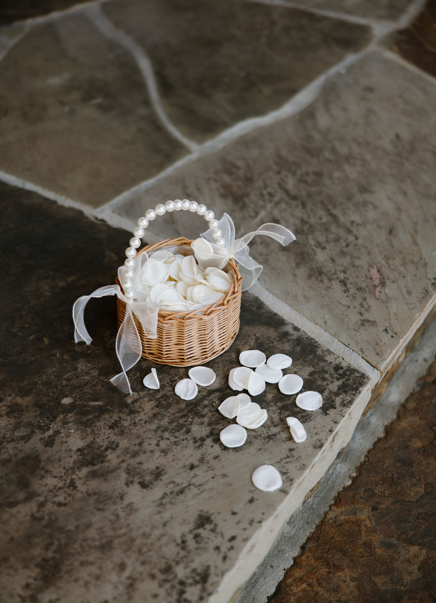 Small wicker flower girl basket with pearl handle and white rose petals on stone floor.
