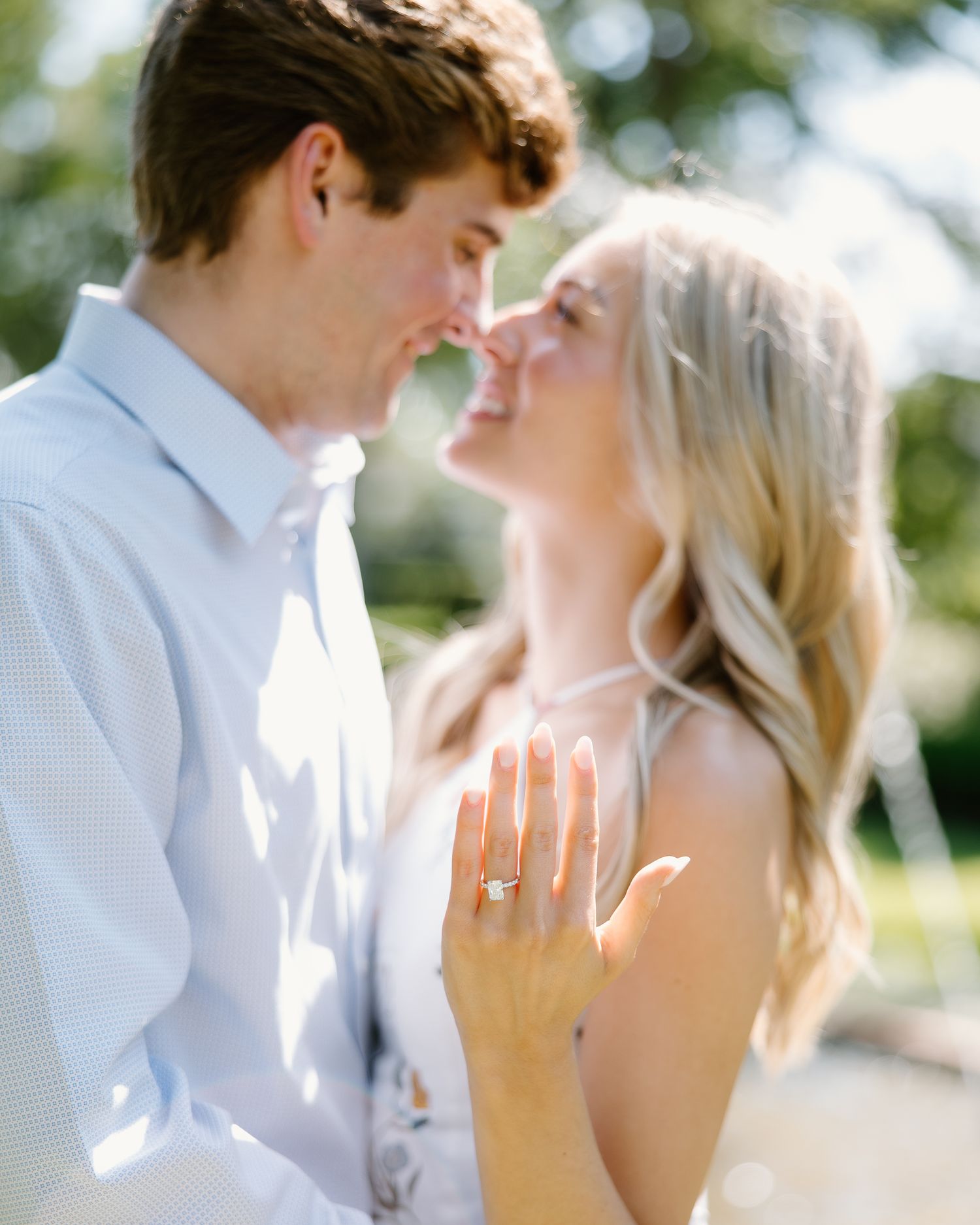 Smiling blonde woman shows engagement ring while leaning close to her partner outdoors.