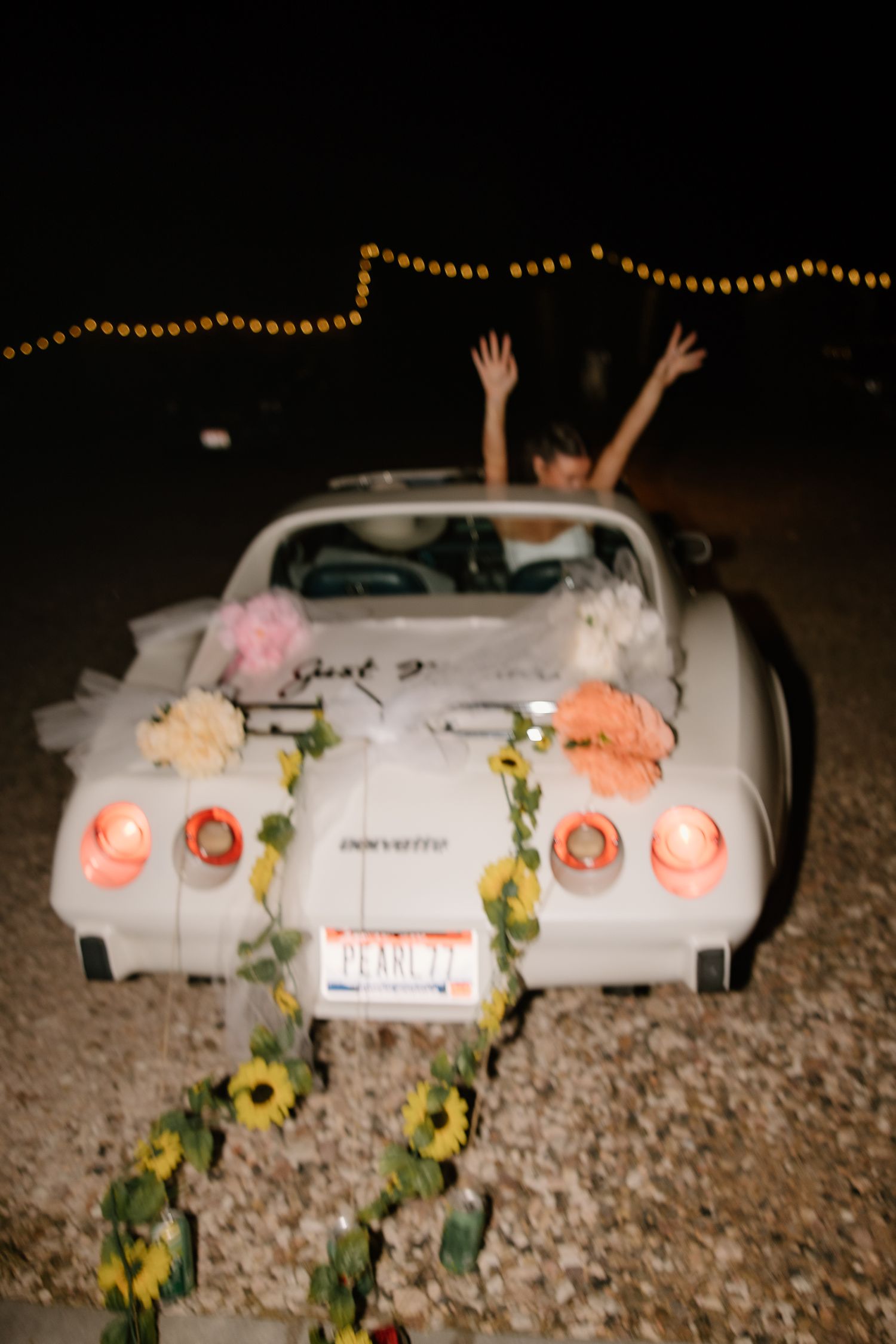 Bride and groom celebrate in decorated white Corvette convertible with flowers at night.