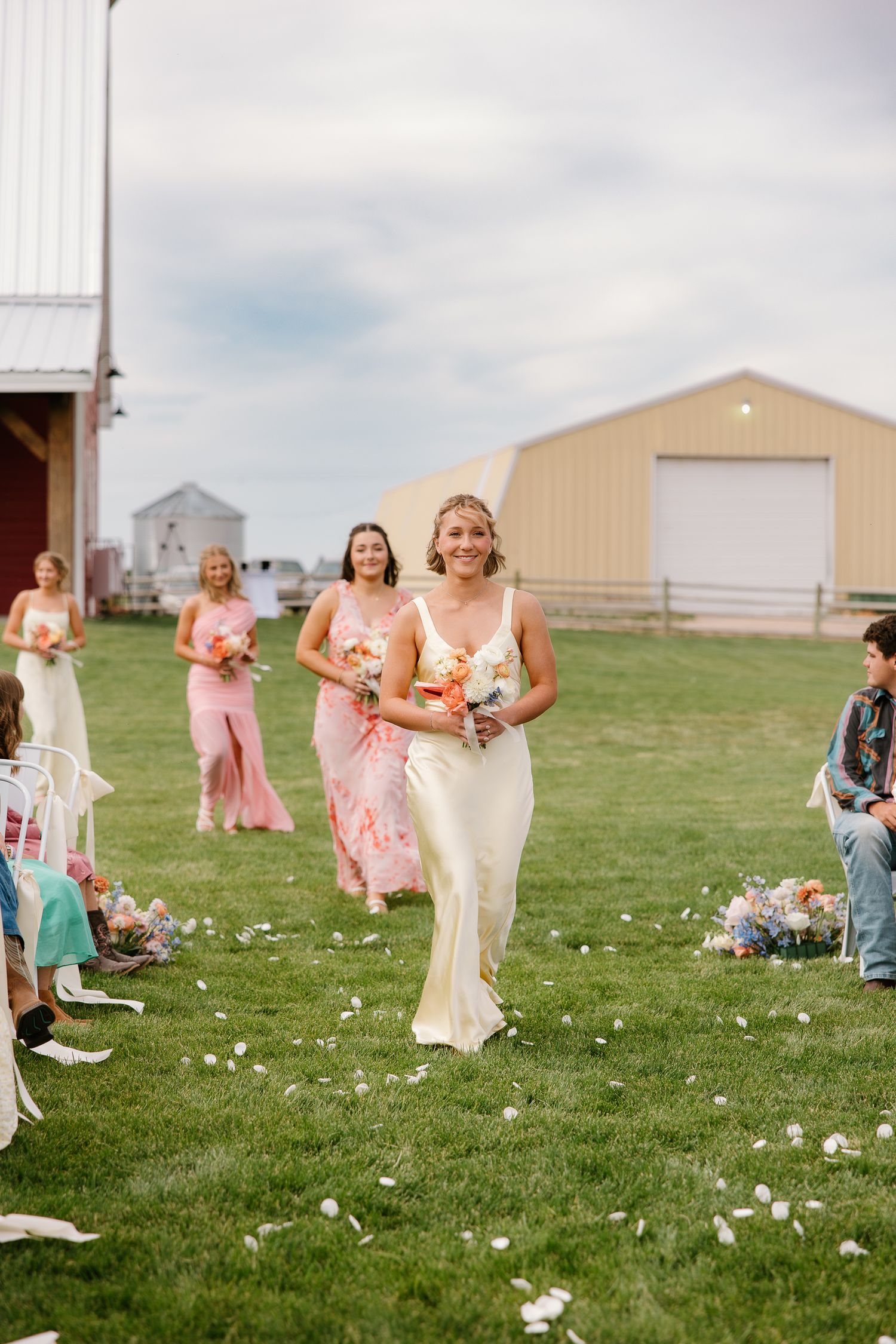 Bride in a white gown walks down the outdoor aisle with bridesmaids in pink dresses at a farm wedding.