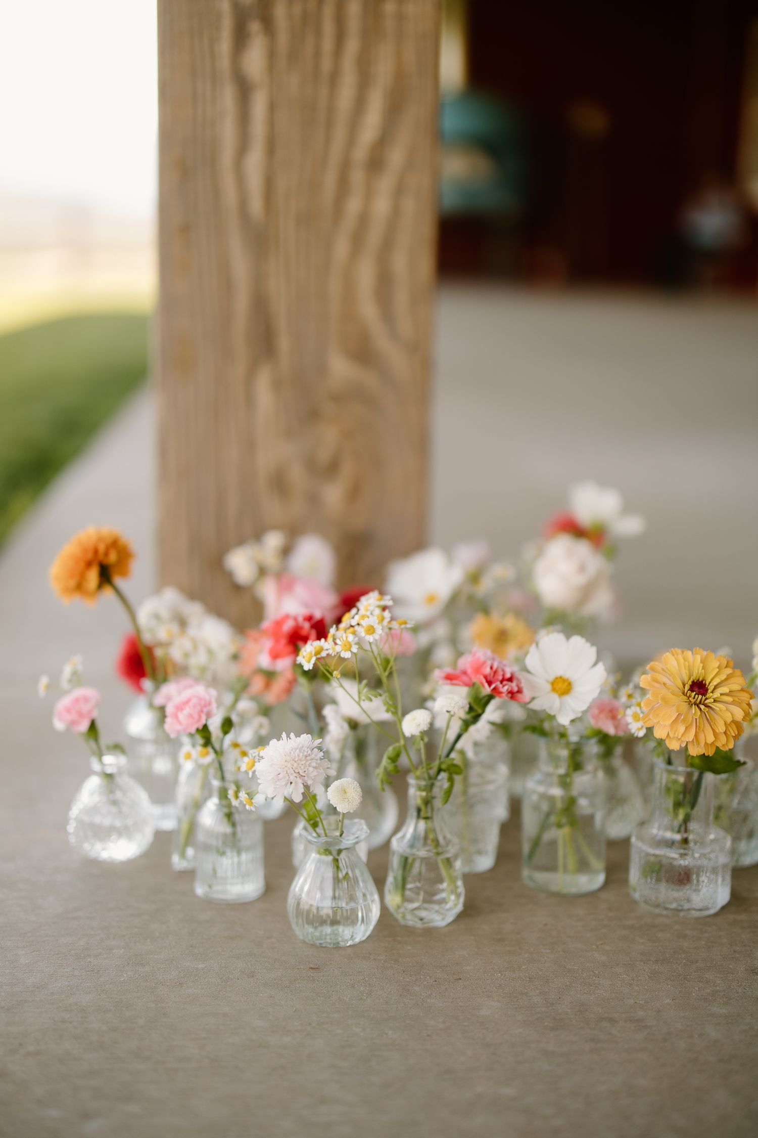 Collection of small glass bud vases with colorful wildflowers arranged on a concrete surface near a wooden post.
