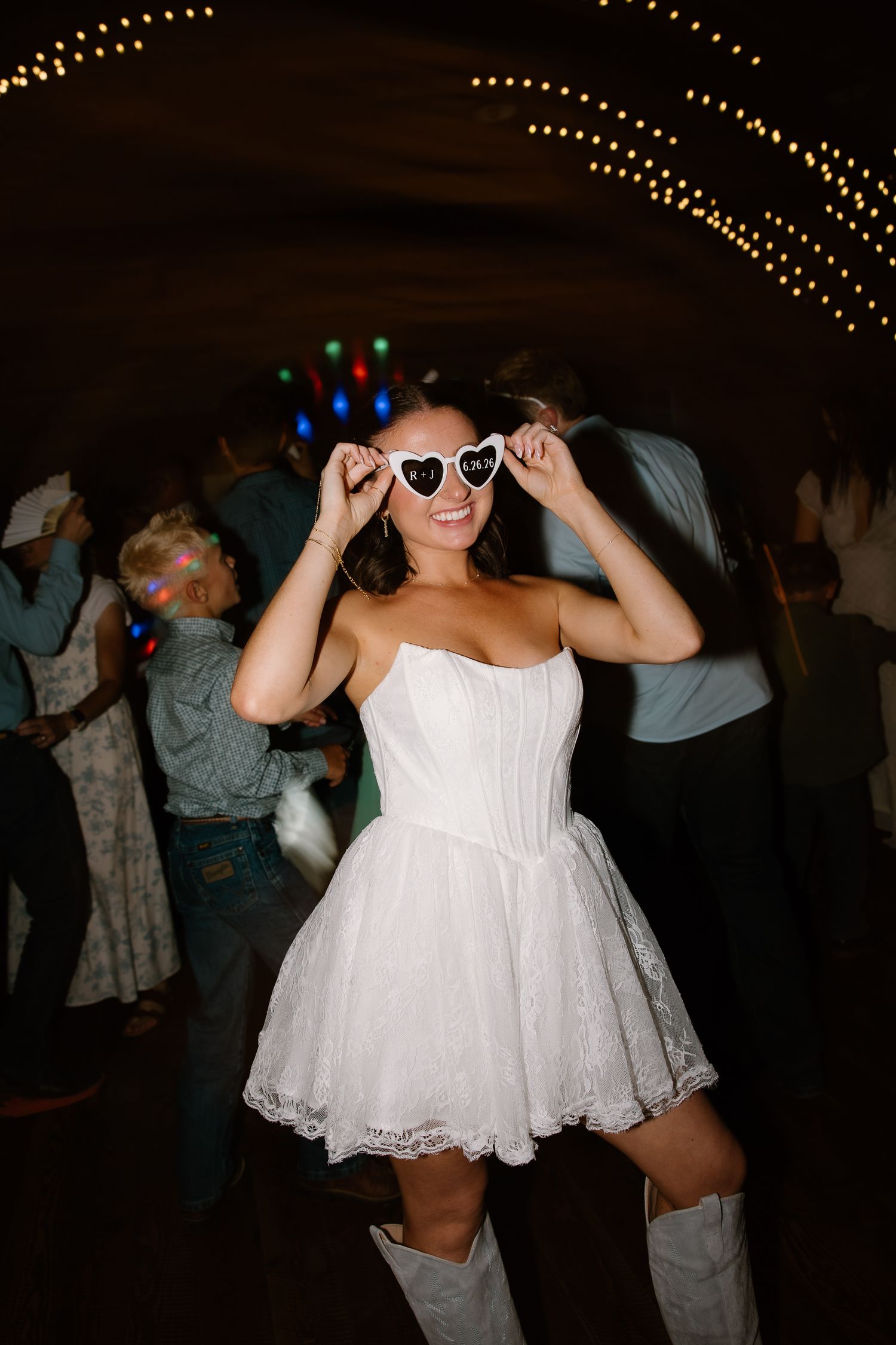 Bride in short white lace dress and heart-shaped sunglasses dancing joyfully at wedding reception.
