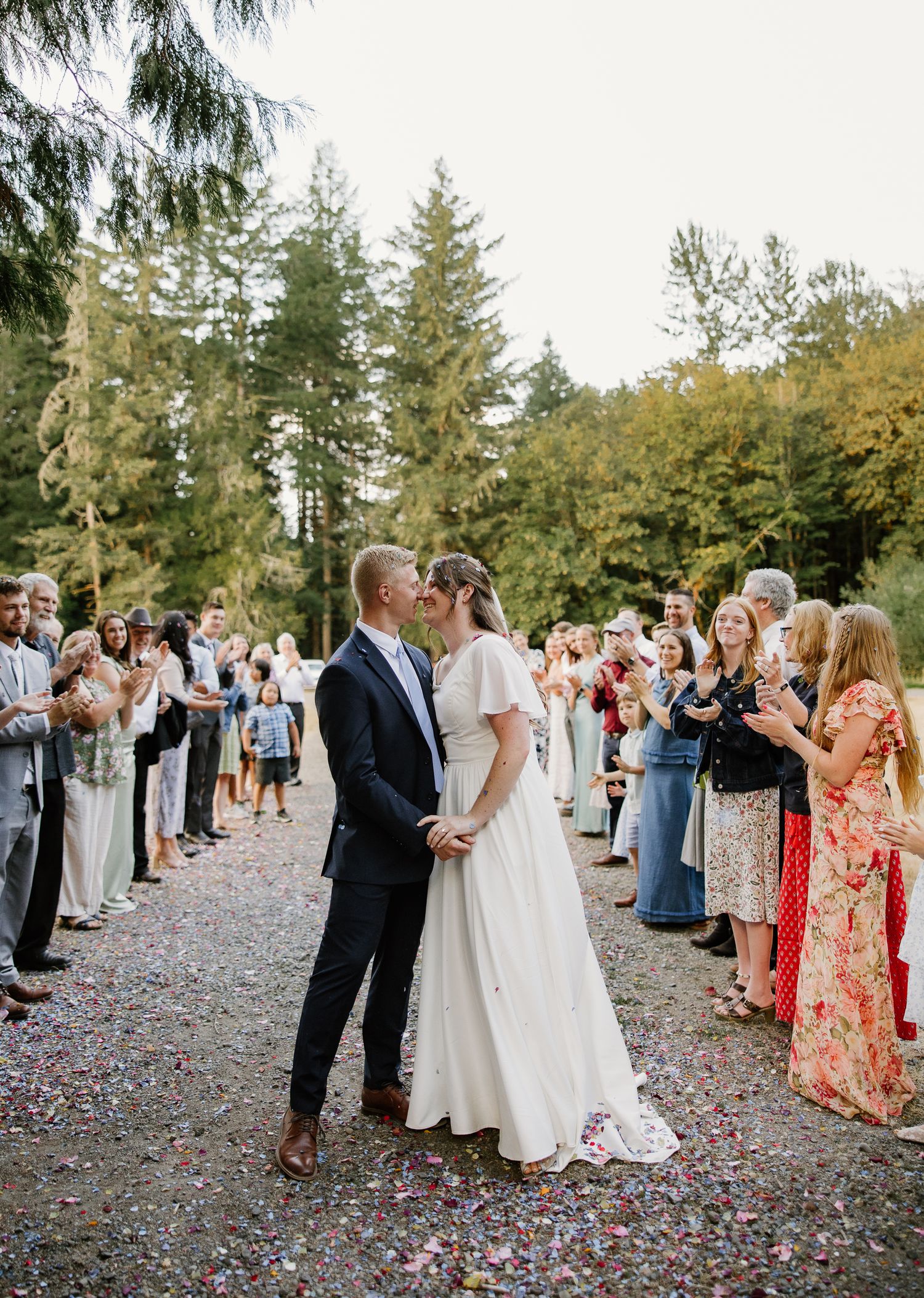 Bride and groom kiss during confetti send-off on tree-lined path surrounded by cheering guests.