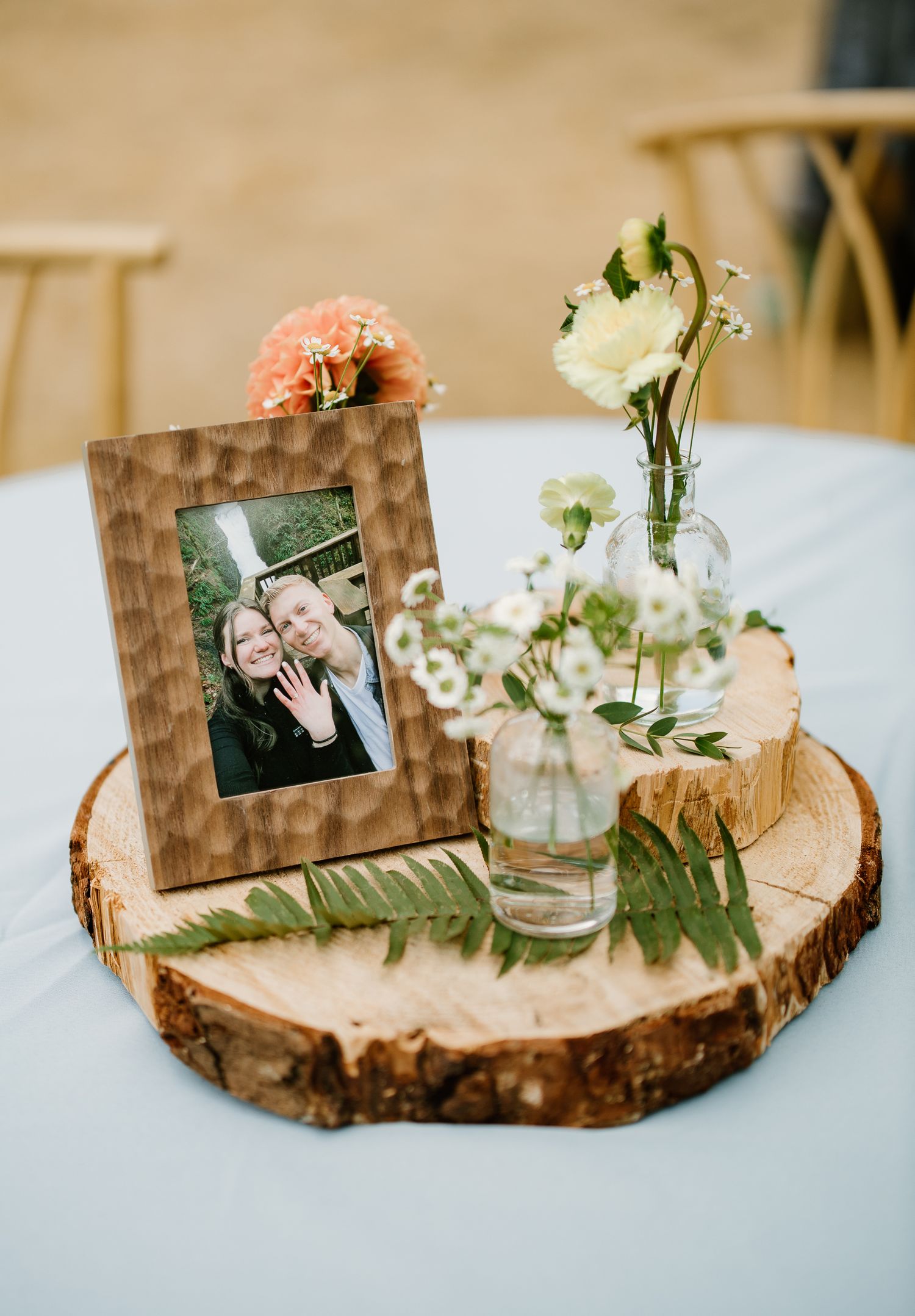 Rustic wedding centerpiece with wood slice, bud vases, wildflowers, ferns, and a framed couple photo.