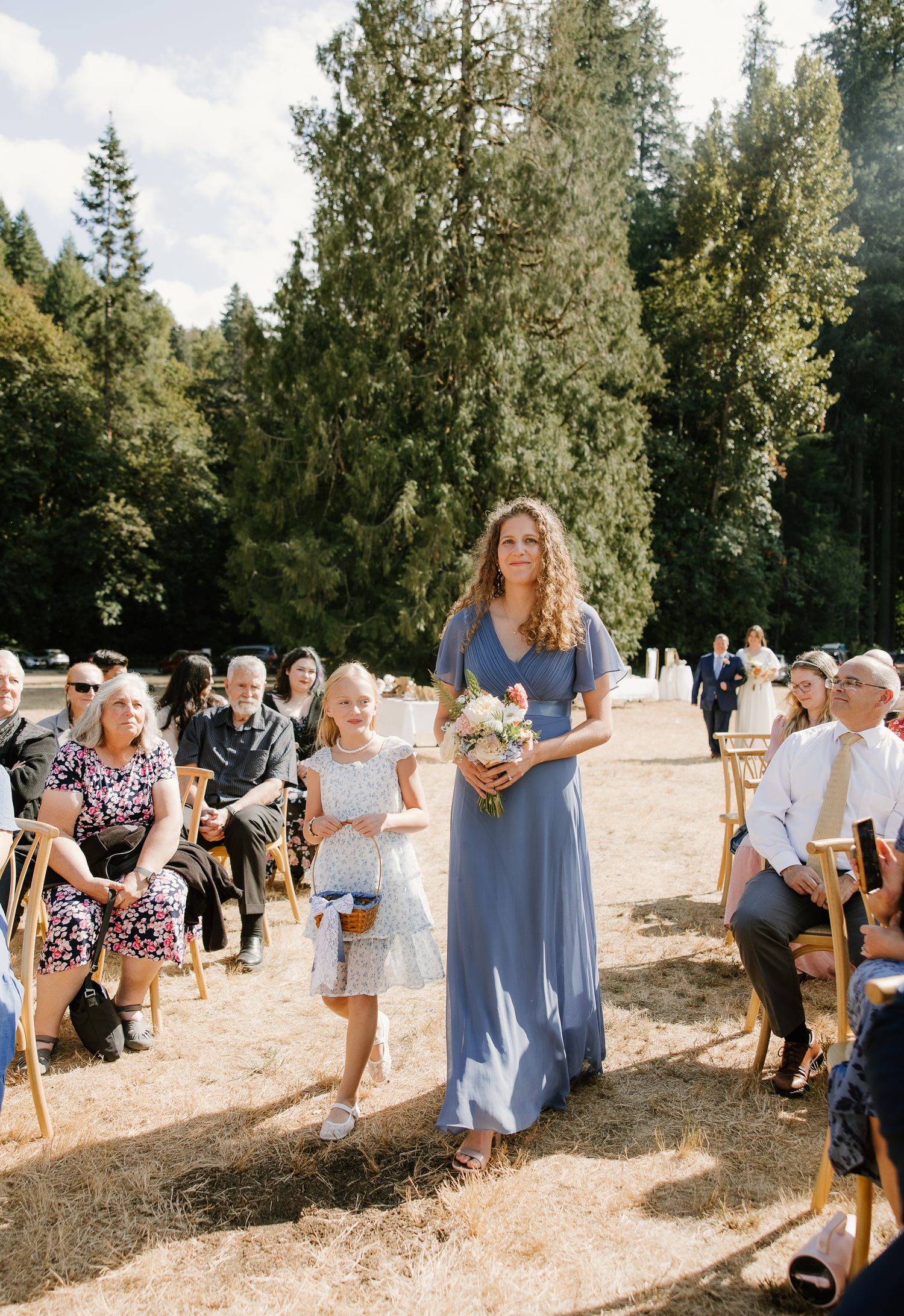 A bridesmaid in a blue dress walks down the aisle with a flower girl at an outdoor wedding ceremony.