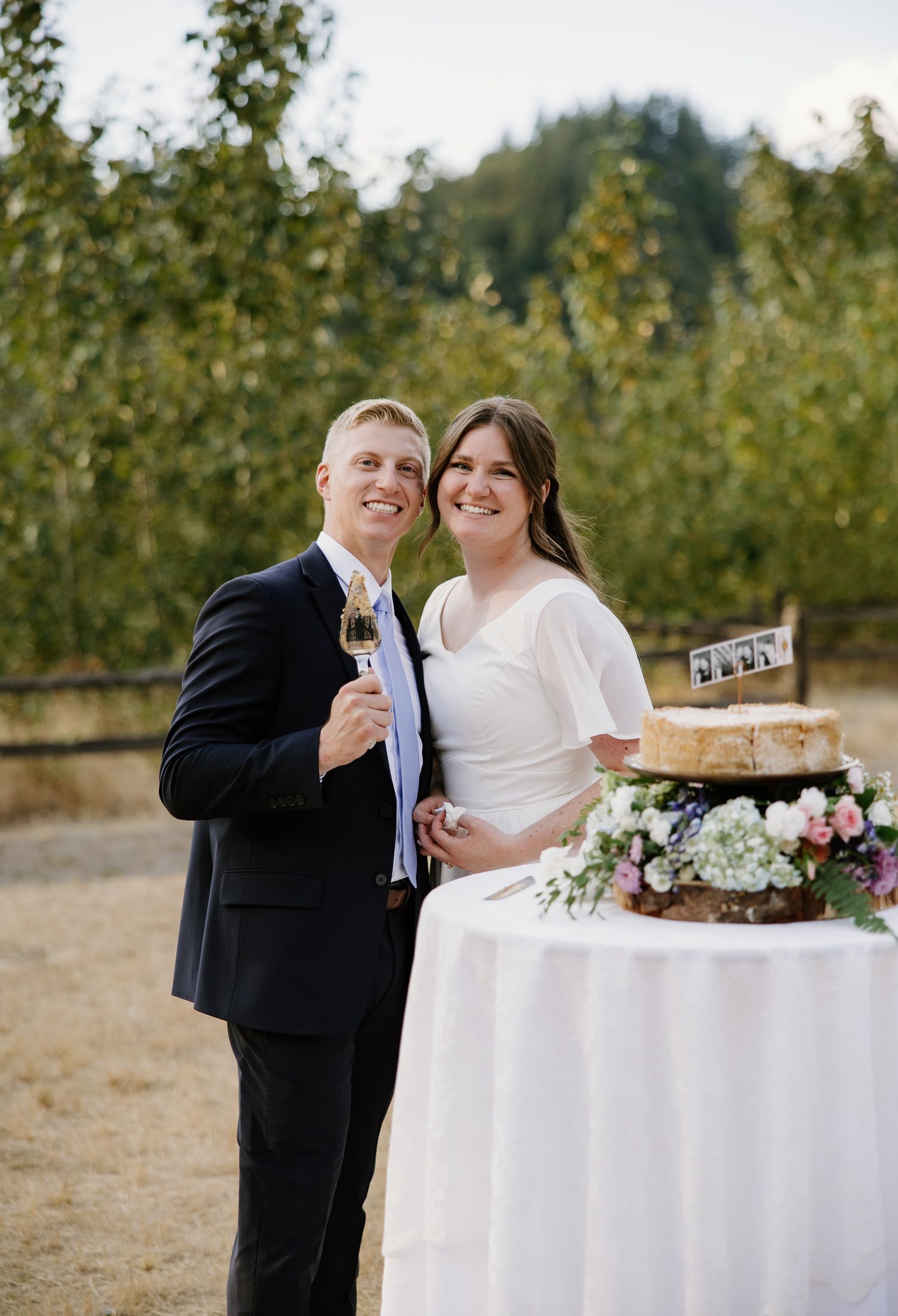 Smiling bride and groom pose beside their wedding cake at an outdoor orchard reception.