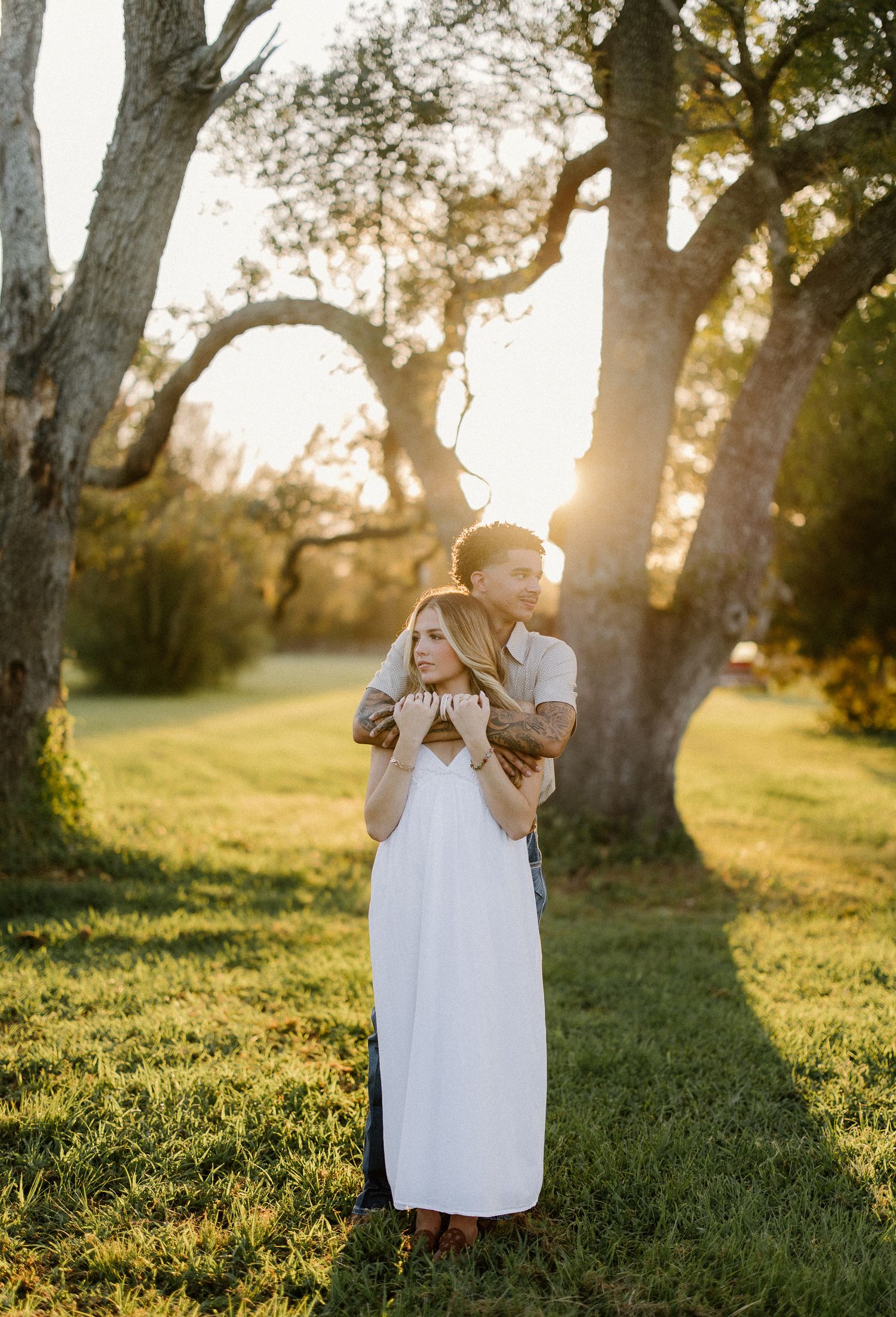 Couple embracing at golden hour in a sunlit park, woman in white maxi dress, man with tattoos, surrounded by oak trees.