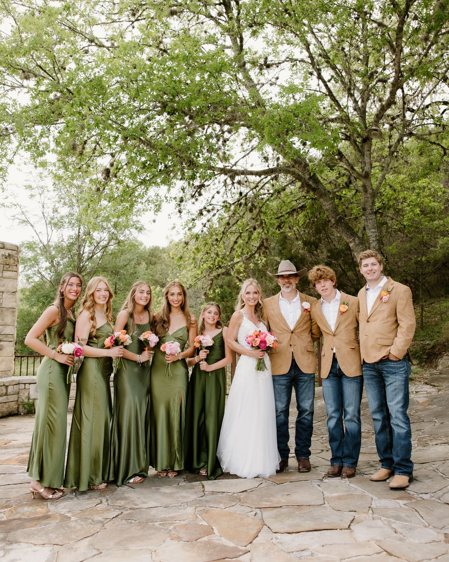 Wedding party poses outdoors, bridesmaids in green gowns, groomsmen in tan blazers and jeans, bride in white gown holding pink bouquet.