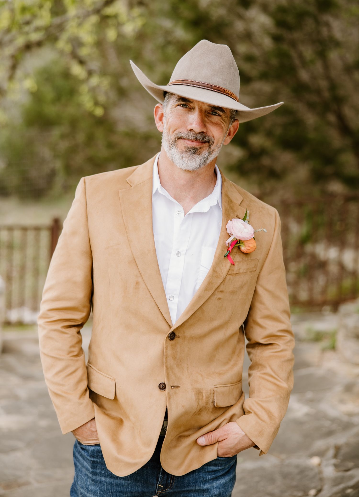 Man in tan blazer, cowboy hat, and boutonniere poses outdoors for a rustic wedding look.