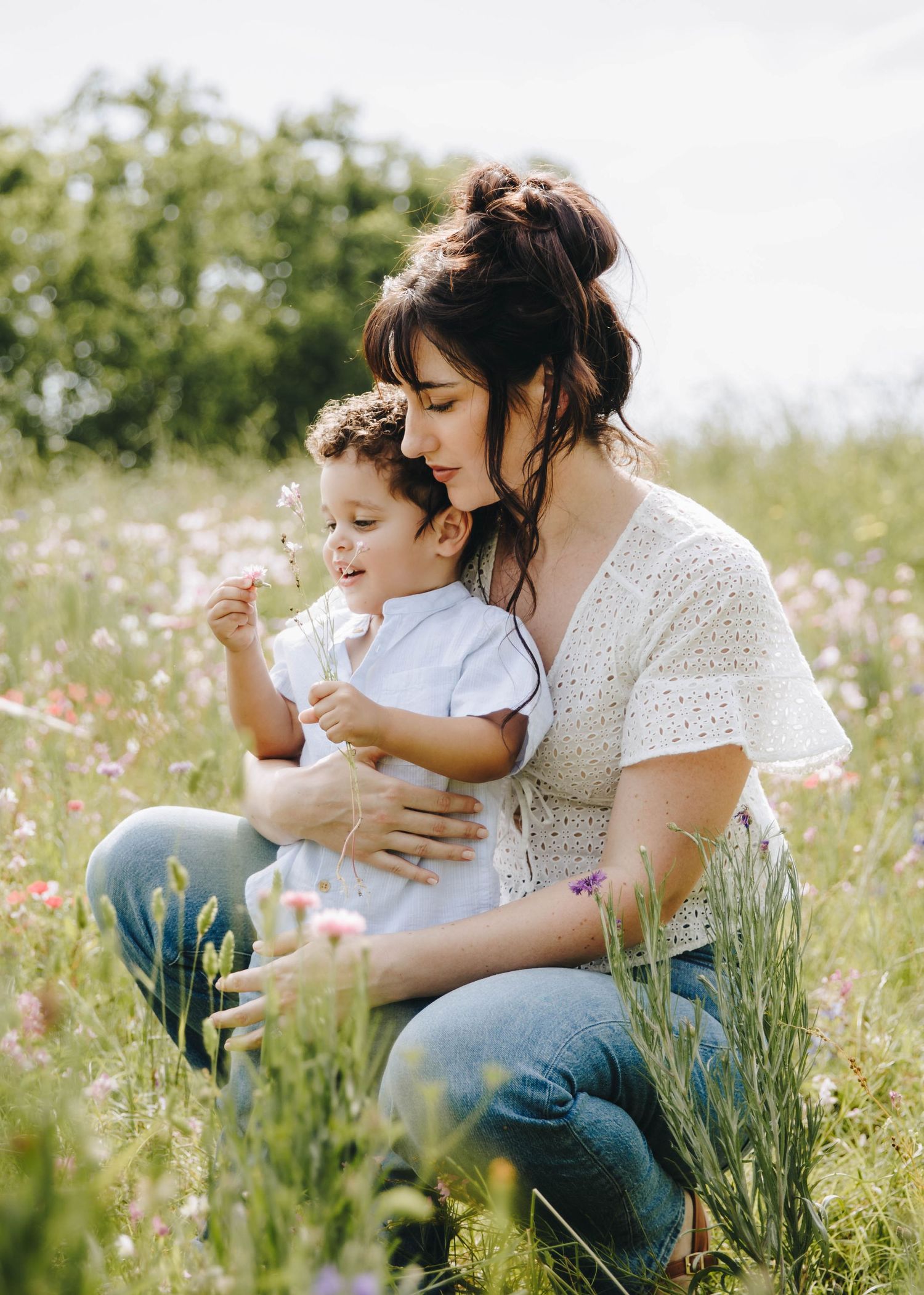 Mother in white lace top gently embraces toddler son while sitting in colorful wildflower field.