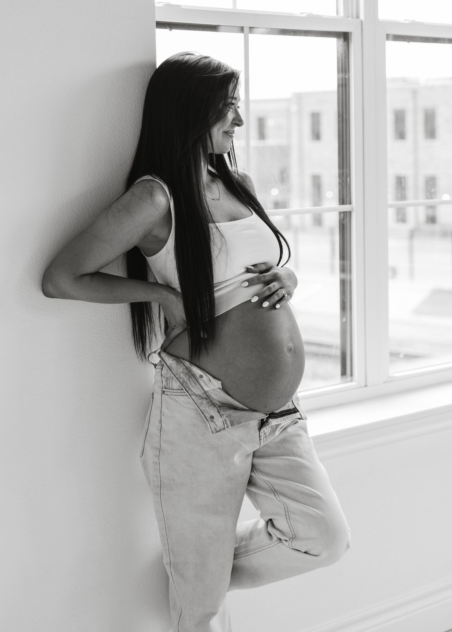 Black and white photo of pregnant woman leaning against window, smiling and holding baby bump.