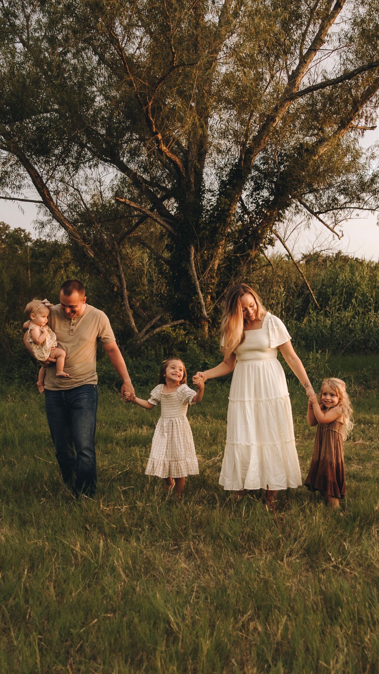 Happy family of five holding hands walking through a lush green meadow during golden hour sunset.
