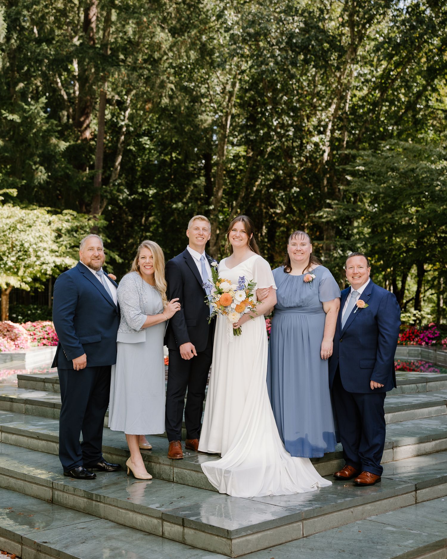 Wedding party of six poses outdoors in formal attire with bride holding colorful bouquet amid lush greenery.