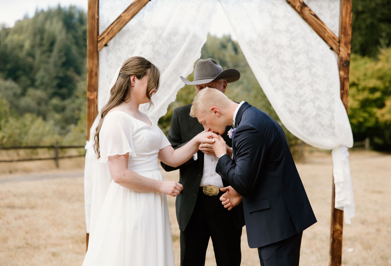 Groom kisses bride's hand during outdoor wedding ceremony under a draped wooden arch.