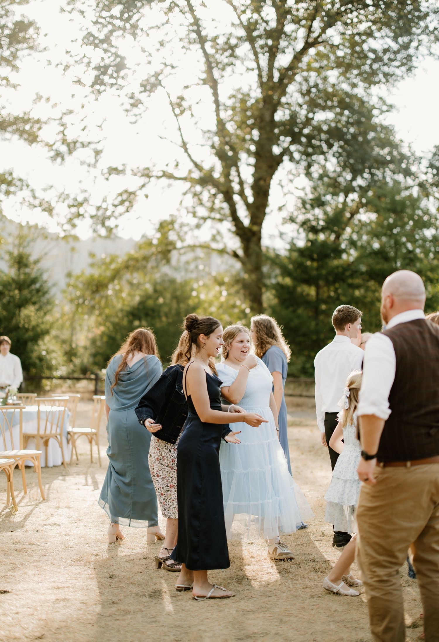 Wedding guests mingle outdoors at a rustic reception venue with tall trees and warm golden sunlight.