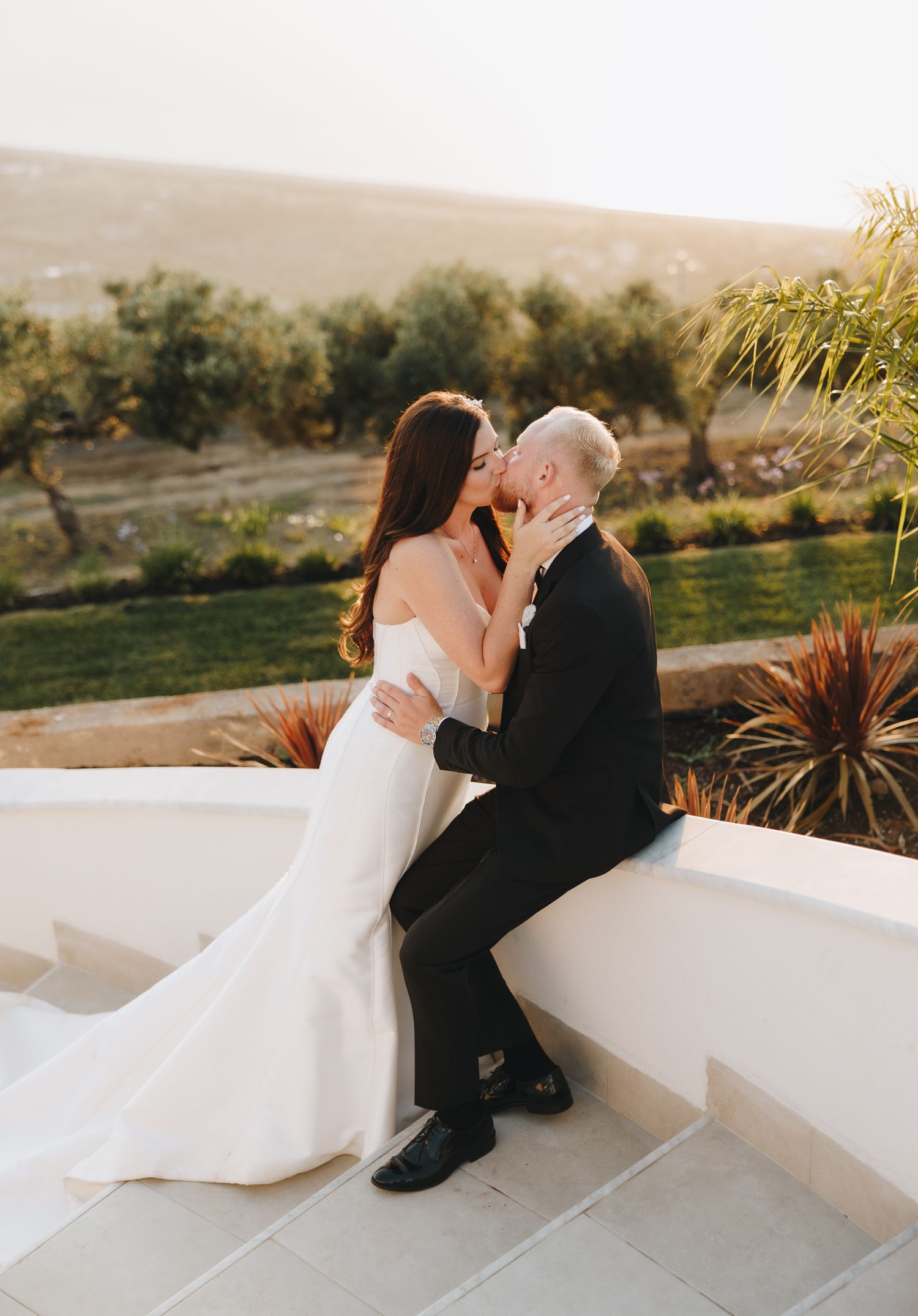 Newlyweds share a romantic kiss on white terrace wall with olive grove backdrop.