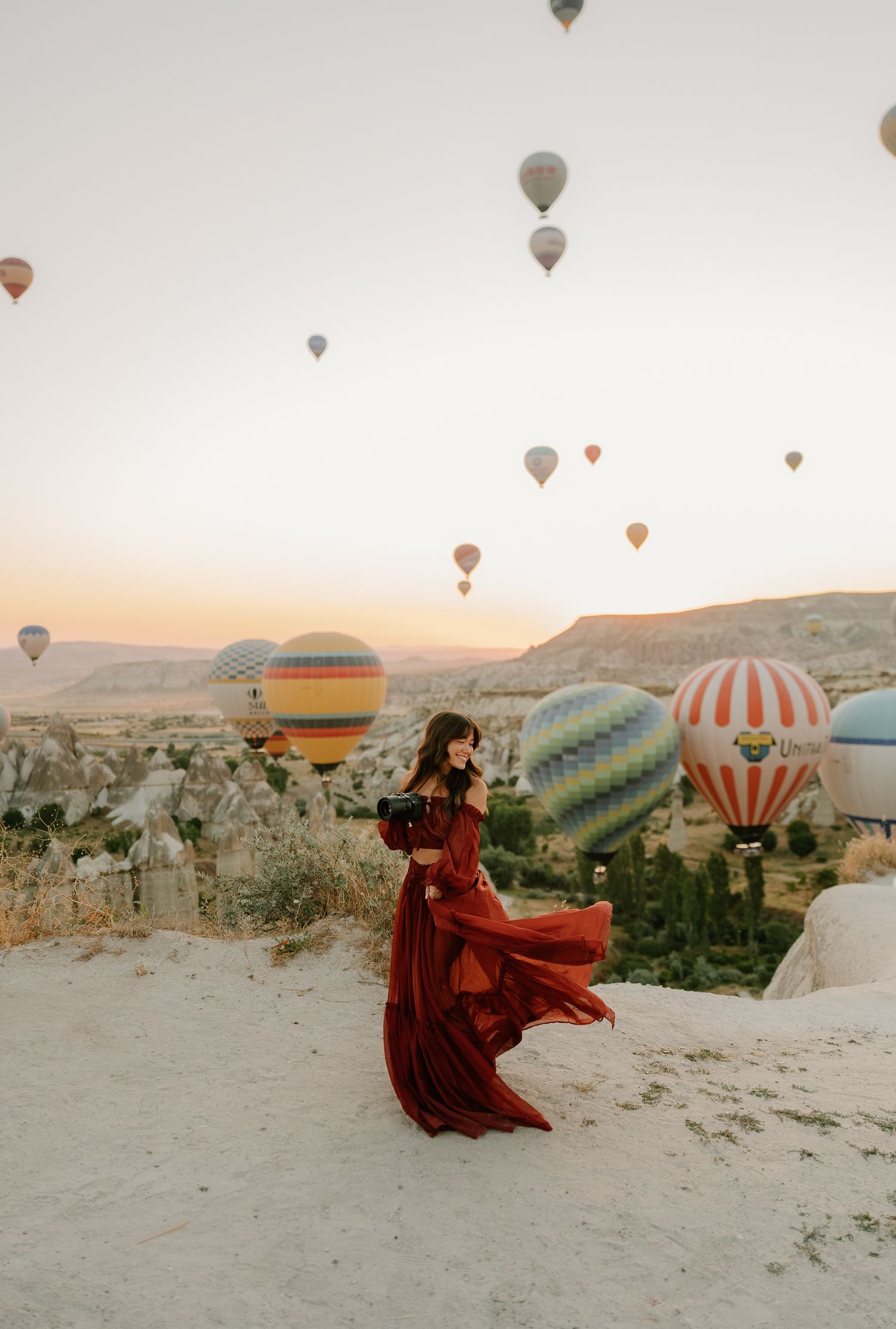 Woman in red flowing dress poses at sunrise in Cappadocia, Turkey, with colorful hot air balloons filling the sky.