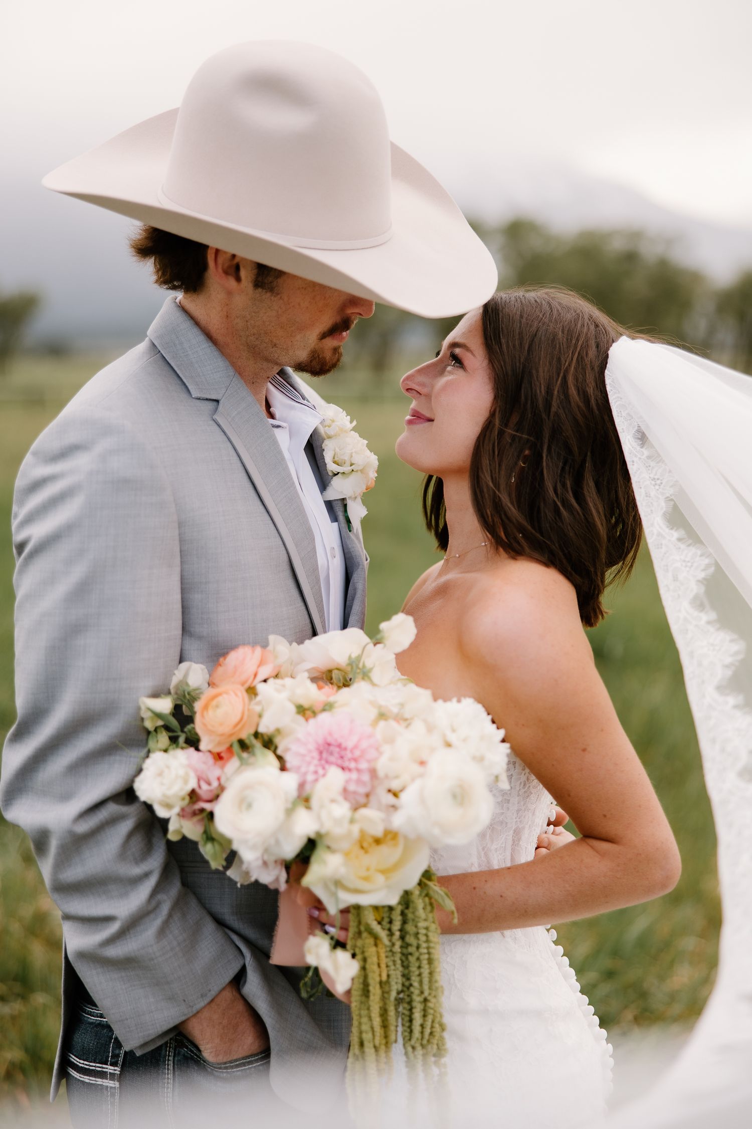 Bride and groom share a romantic moment outdoors, groom in gray suit and cowboy hat, bride holding bouquet.