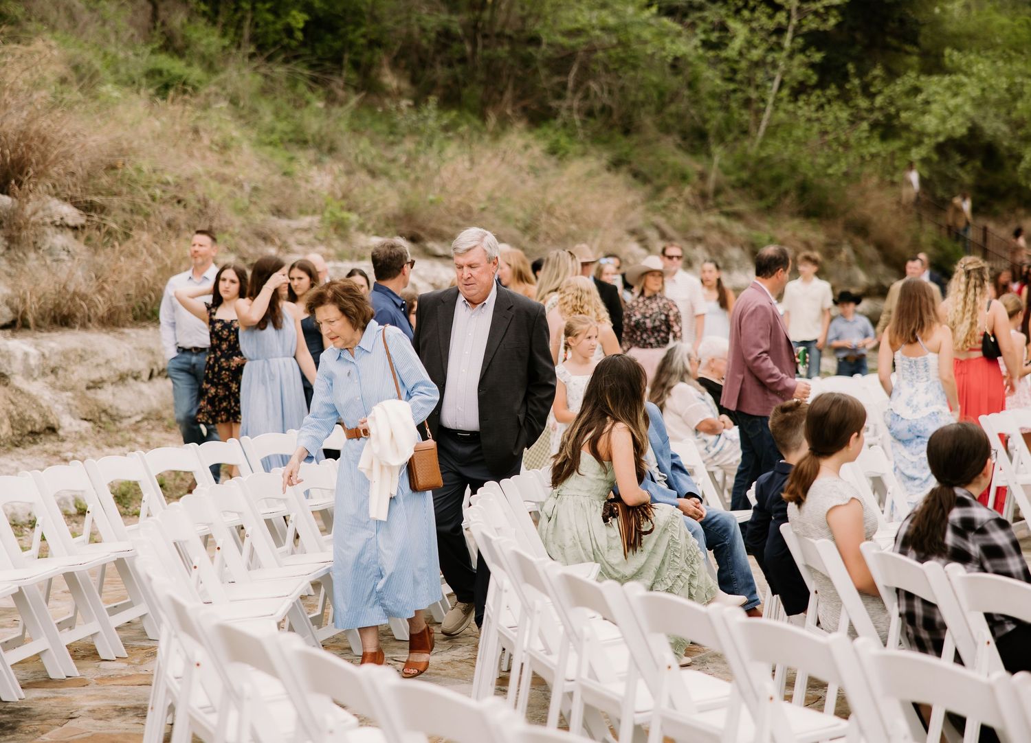 Wedding guests walk between white chairs at an outdoor ceremony venue with rocky terrain.