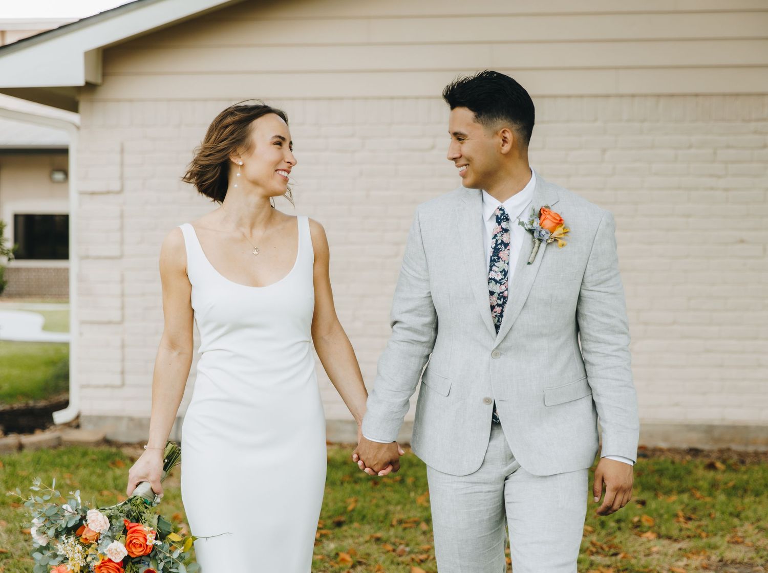 Smiling bride in sleek white dress and groom in light gray suit walk hand in hand holding colorful bouquet.