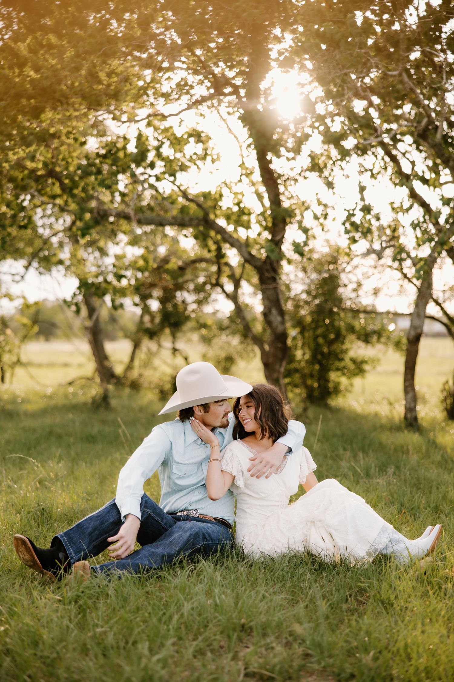 Couple sitting in golden sunlight on grass, man in cowboy hat leaning toward woman in white dress.