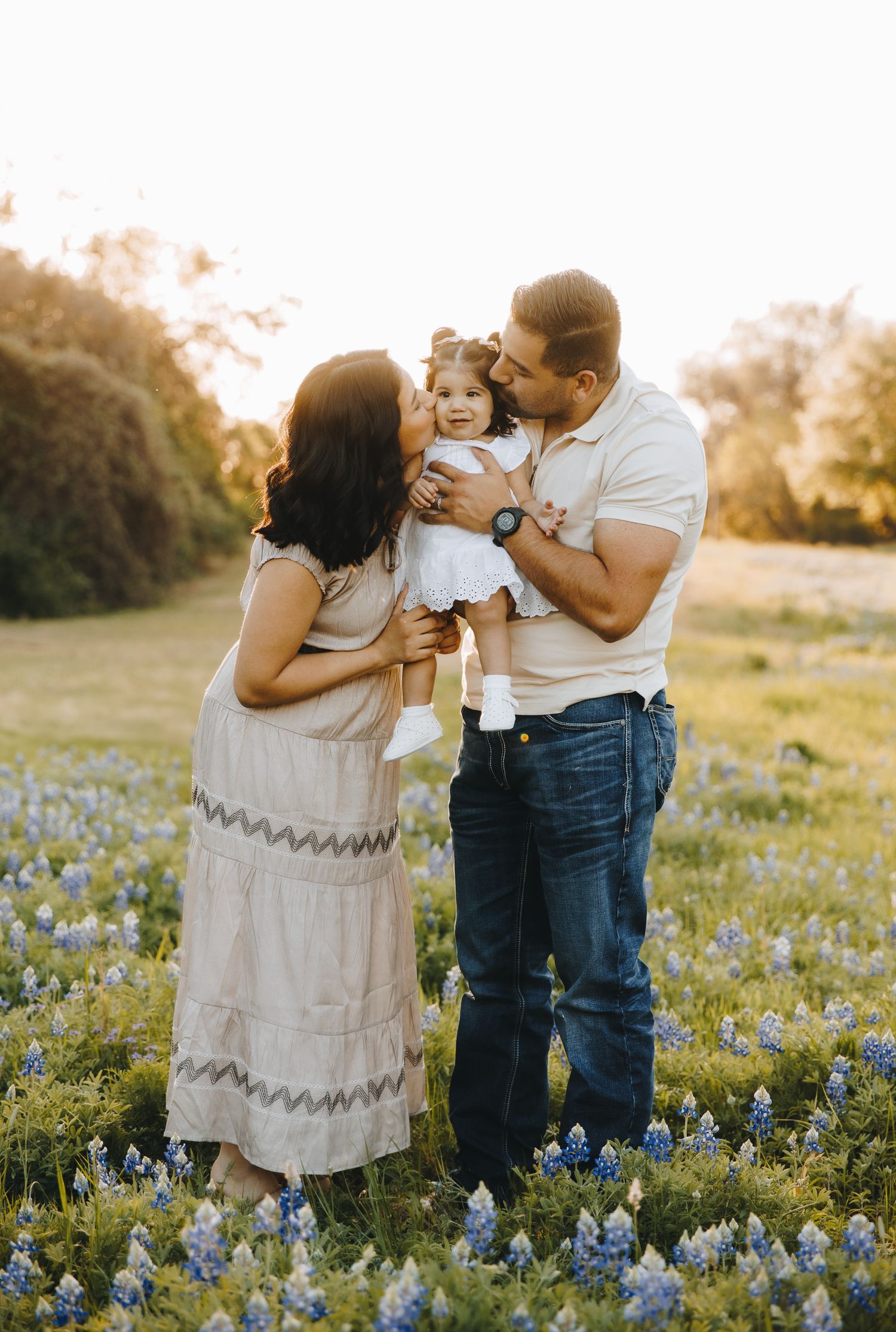 Family portrait in bluebonnet field at golden hour, parents kissing toddler girl in white dress.