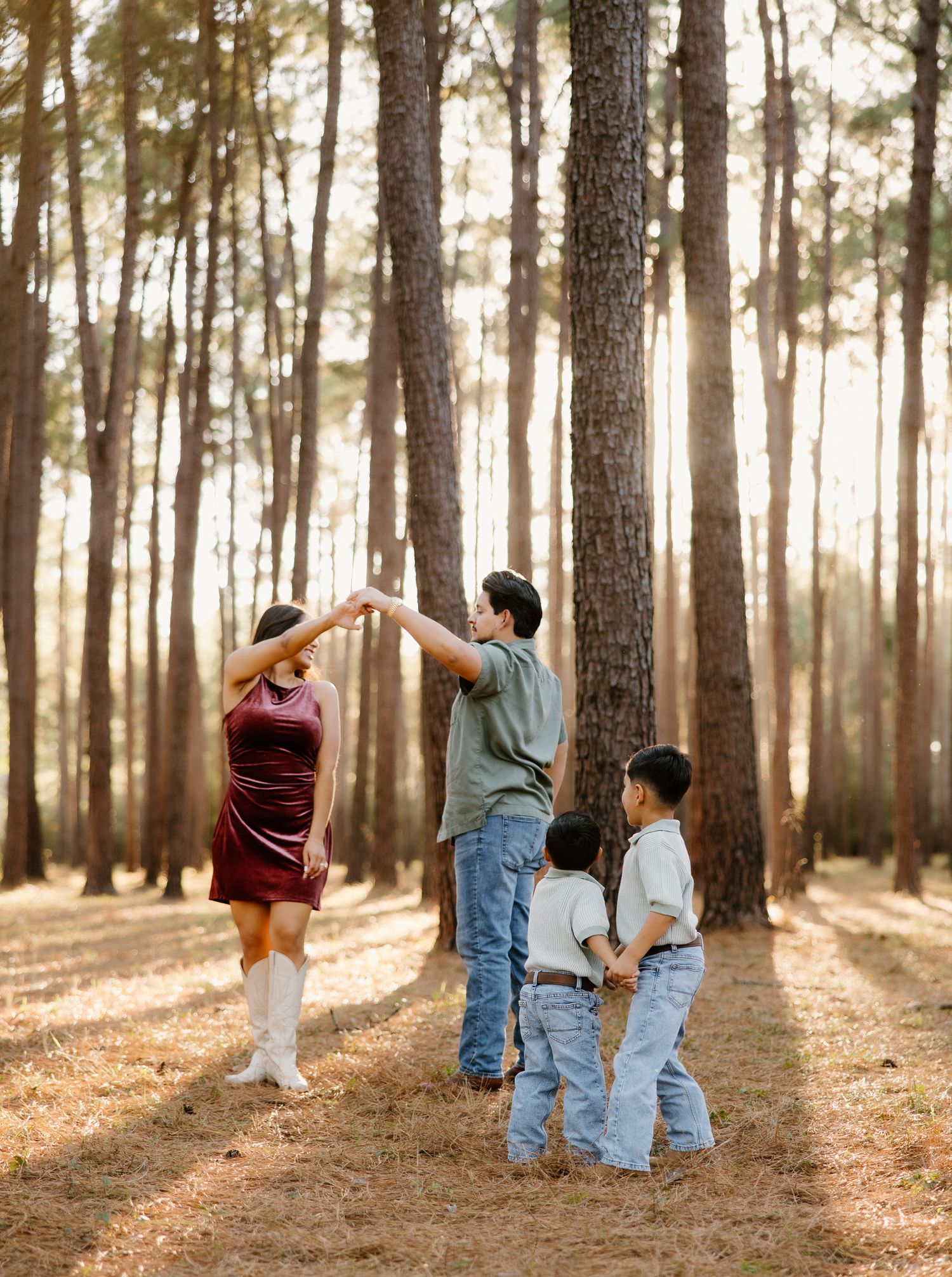 Family of four dances together in sunlit pine forest, parents spinning while two young children watch.