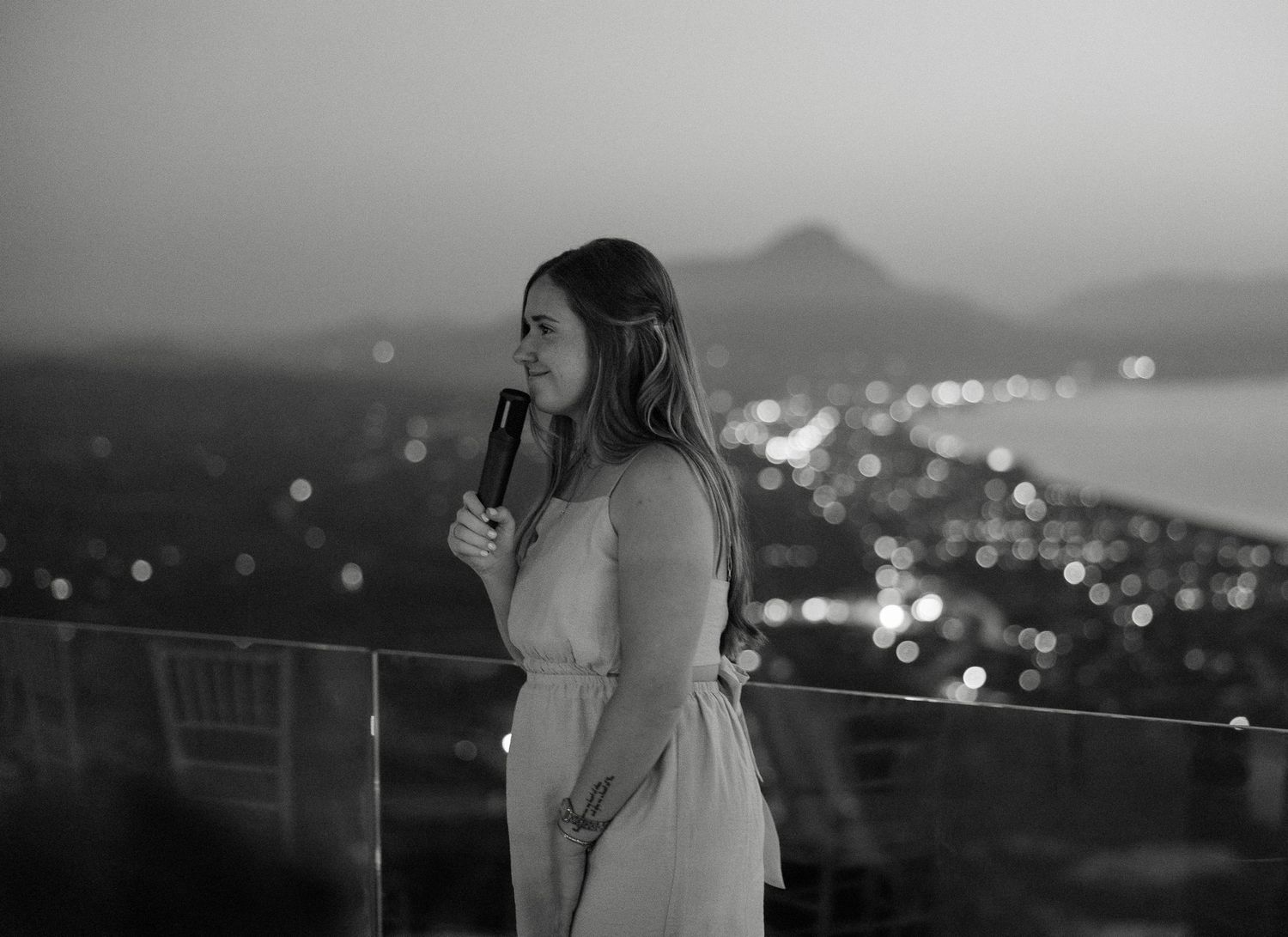 Black and white portrait of a woman speaking into a microphone at a twilight outdoor event.