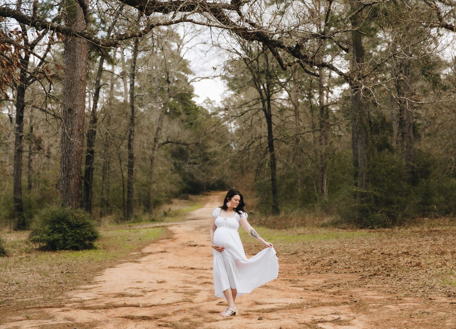 Pregnant woman in white dress twirling on a wooded dirt path during outdoor maternity photo session.