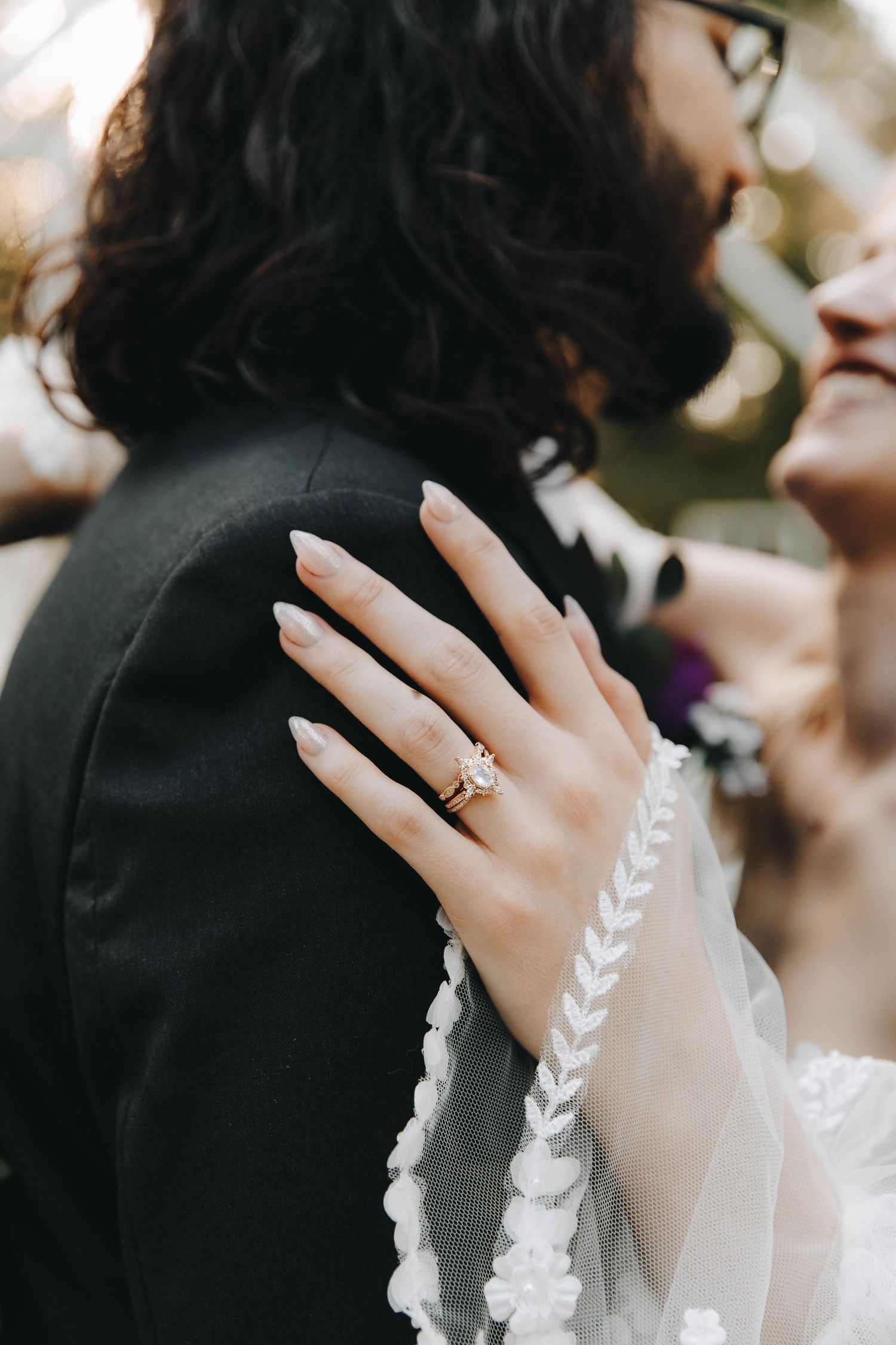 Close-up of bride's hand with a delicate gold engagement ring resting on groom's black suit jacket shoulder.