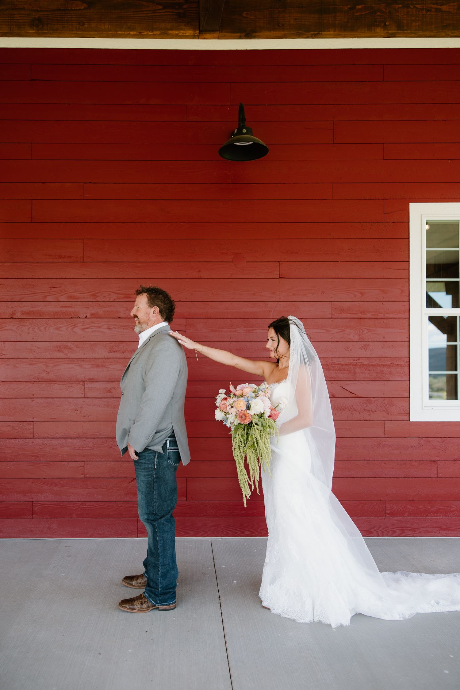 Bride taps groom's shoulder during first look against a red barn wall.
