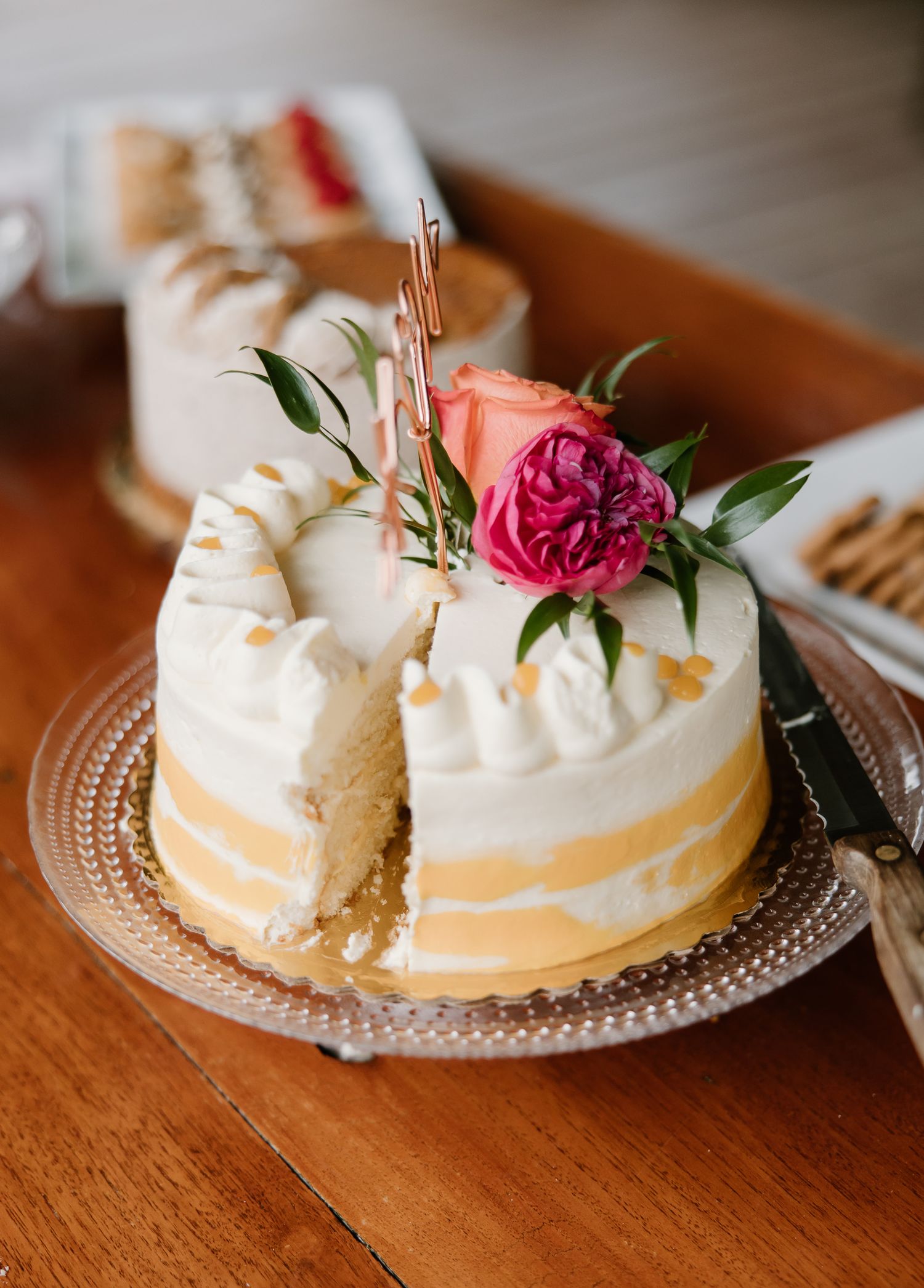 Elegant single-tier white wedding cake with gold accents topped with a pink rose and greenery on a glass plate.