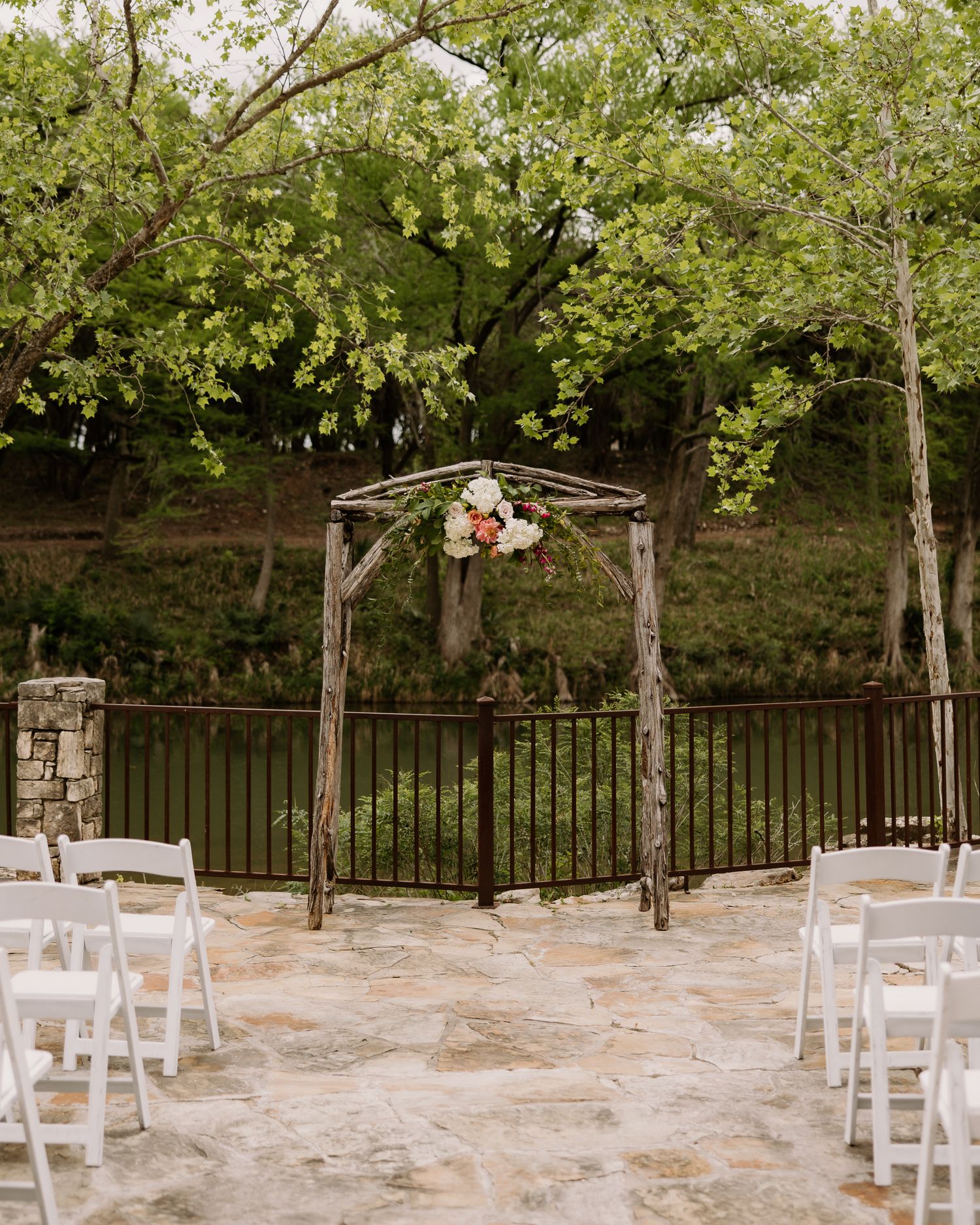 Rustic wooden arch adorned with pink and white flowers at an outdoor wedding ceremony with white chairs on a stone patio.