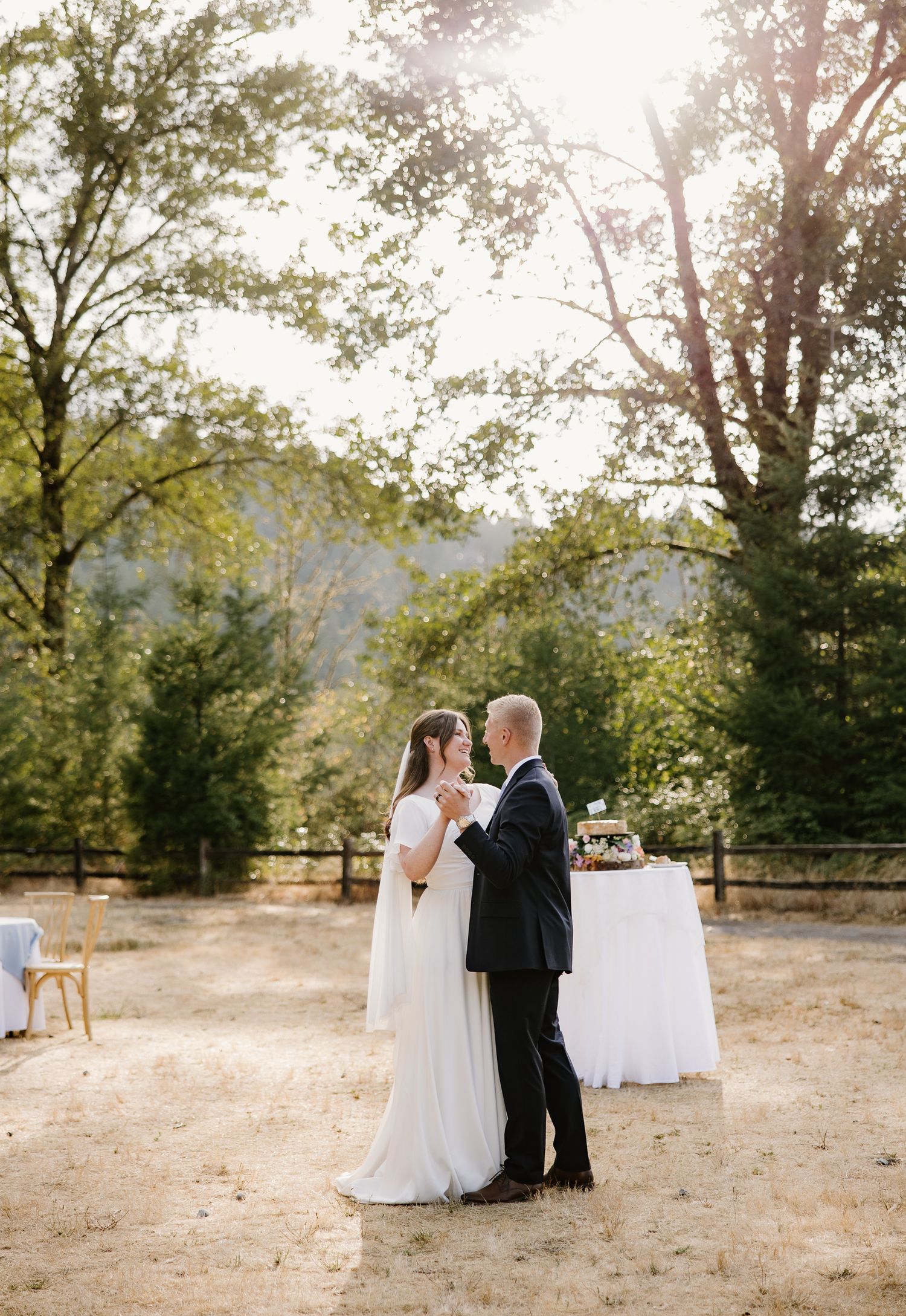 Bride and groom share first dance outdoors at golden hour with sunlit trees and mountains in background.