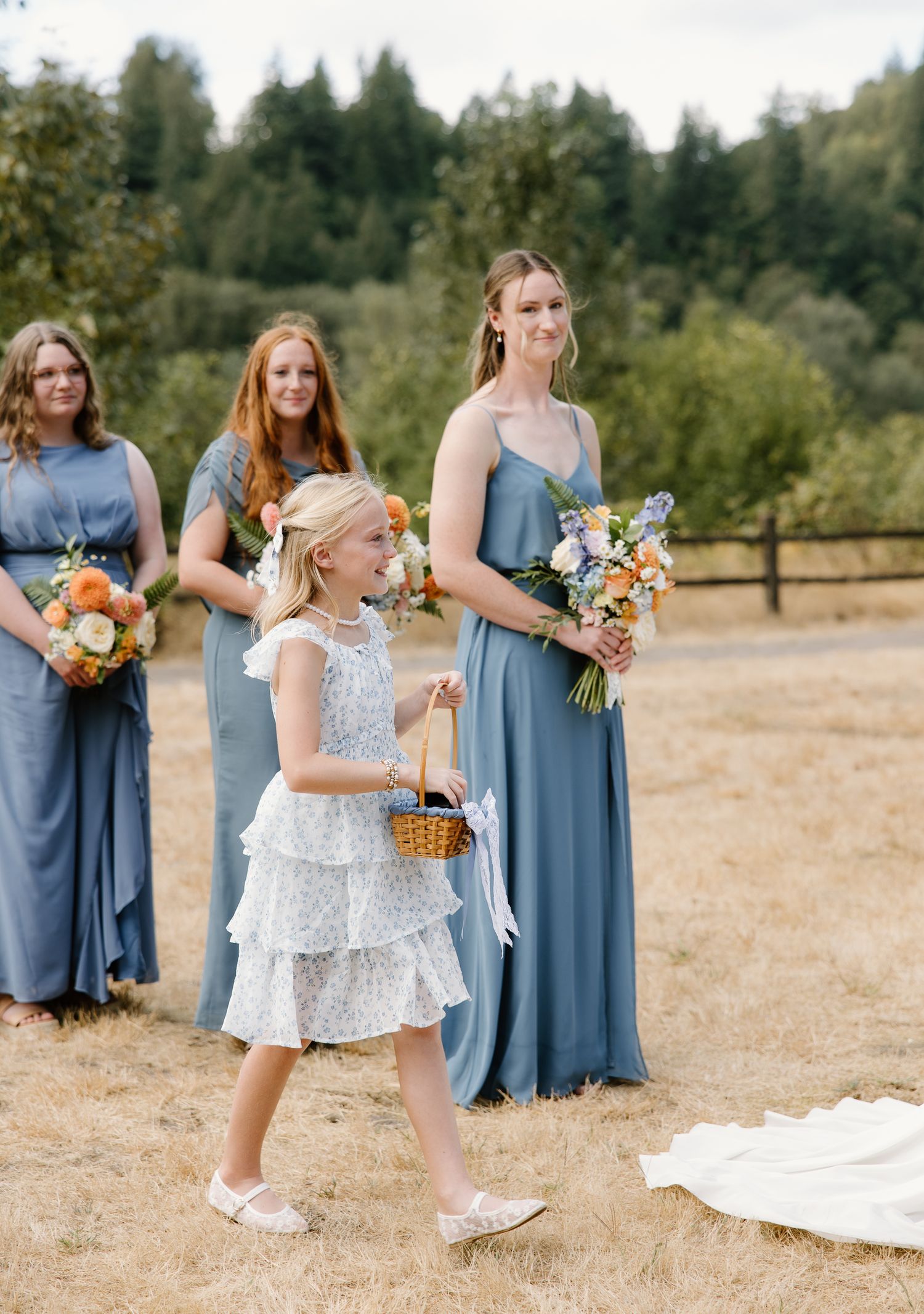 Flower girl in white dress carries basket down aisle as bridesmaids in blue gowns hold colorful bouquets.