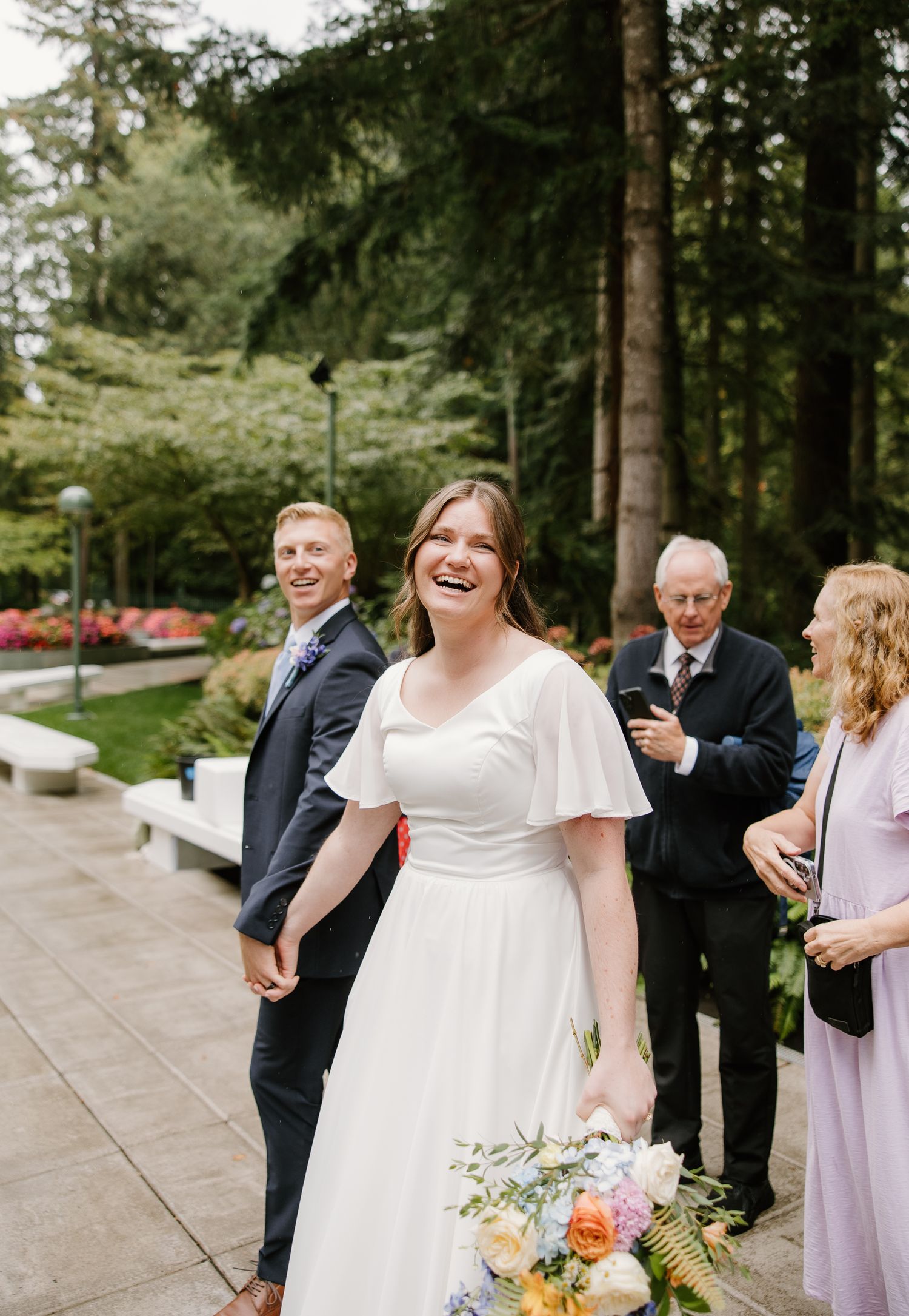 Bride in white flutter-sleeve gown laughing outdoors at garden wedding venue with groom and guests in background.