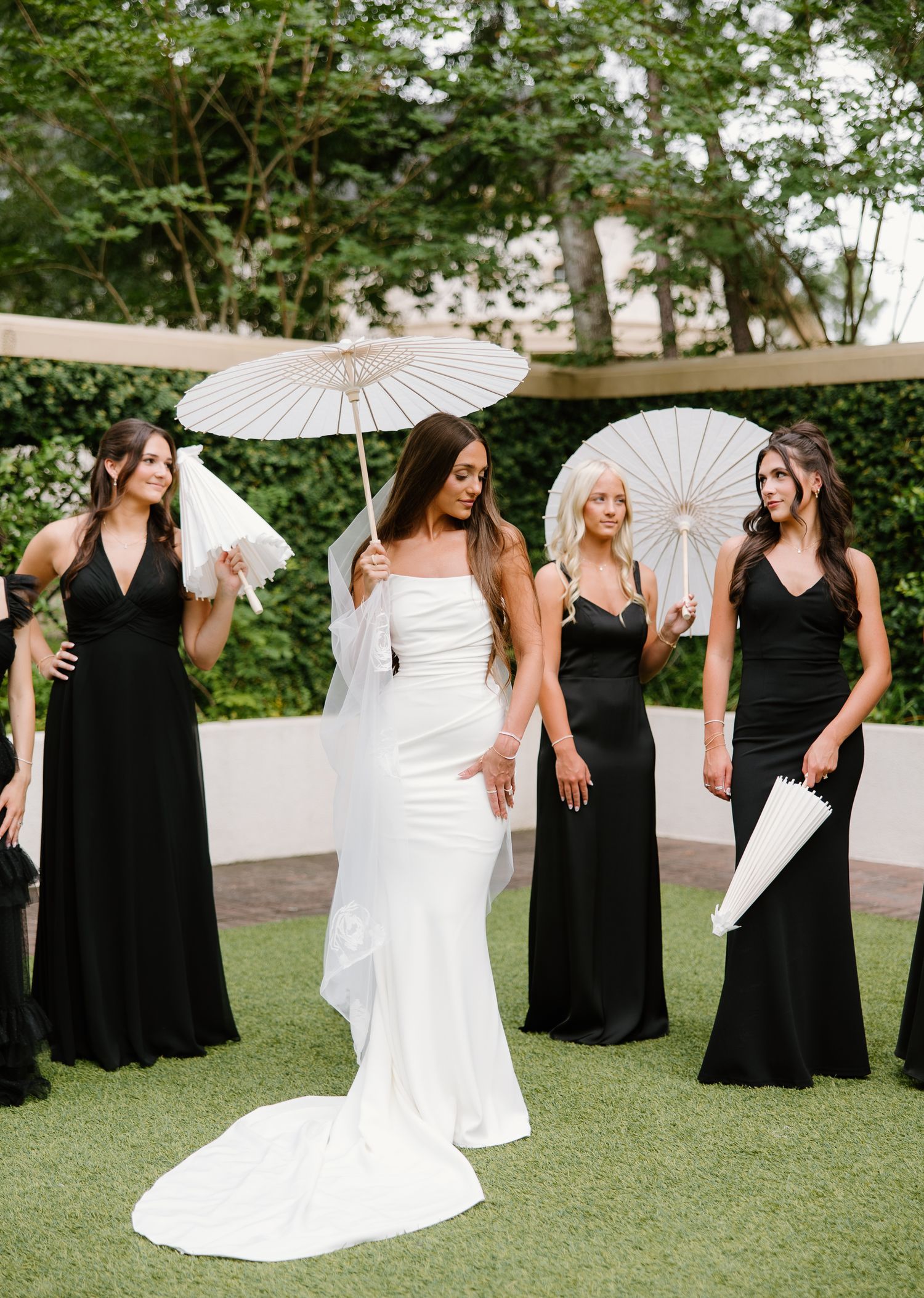 Bride in white gown with bridesmaids in black dresses holding white parasols outdoors on green lawn.