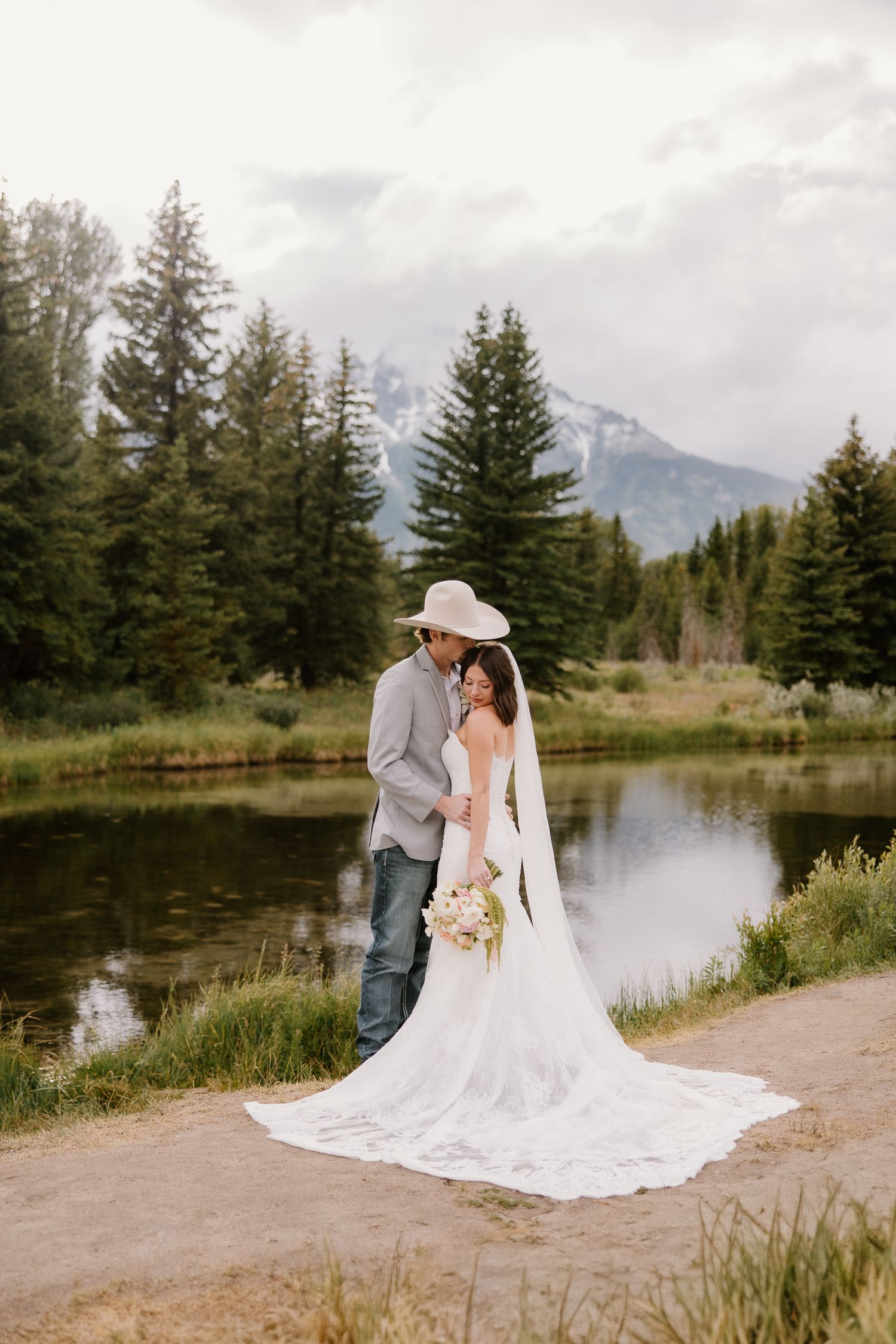 Bride and groom posing by a mountain lake, surrounded by pine trees, bride in white gown with long veil, groom in cowboy hat.