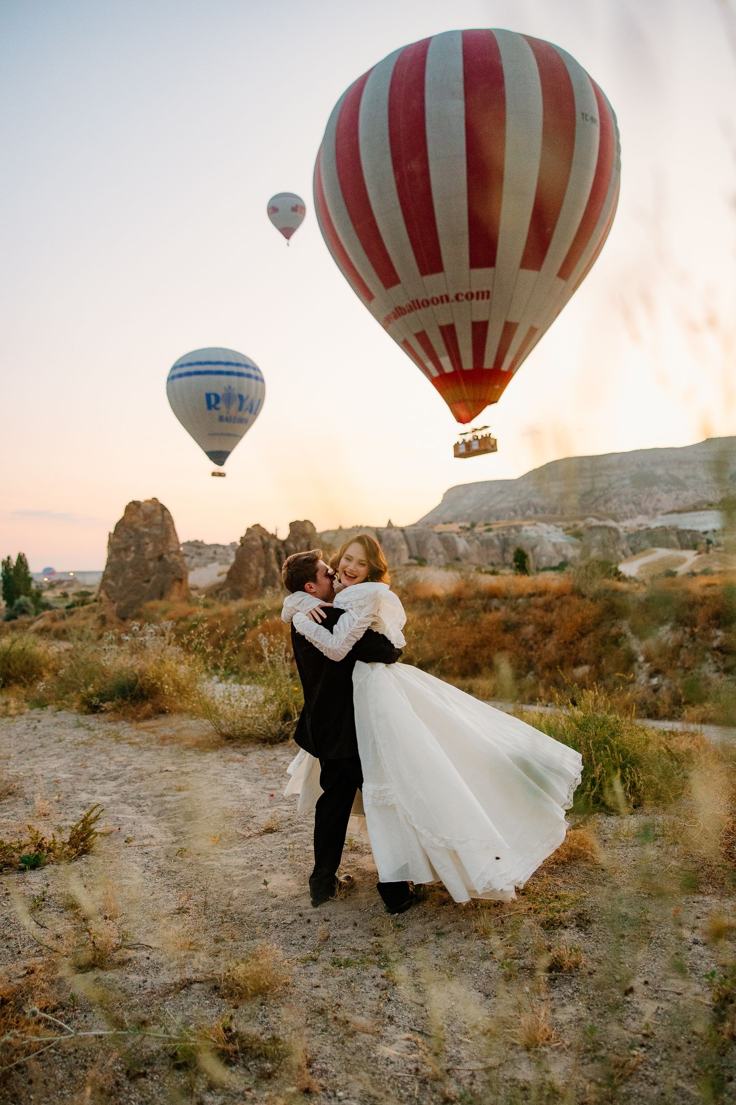 Couple embracing in wedding attire with colorful hot air balloons floating over rocky Cappadocia landscape at sunset.
