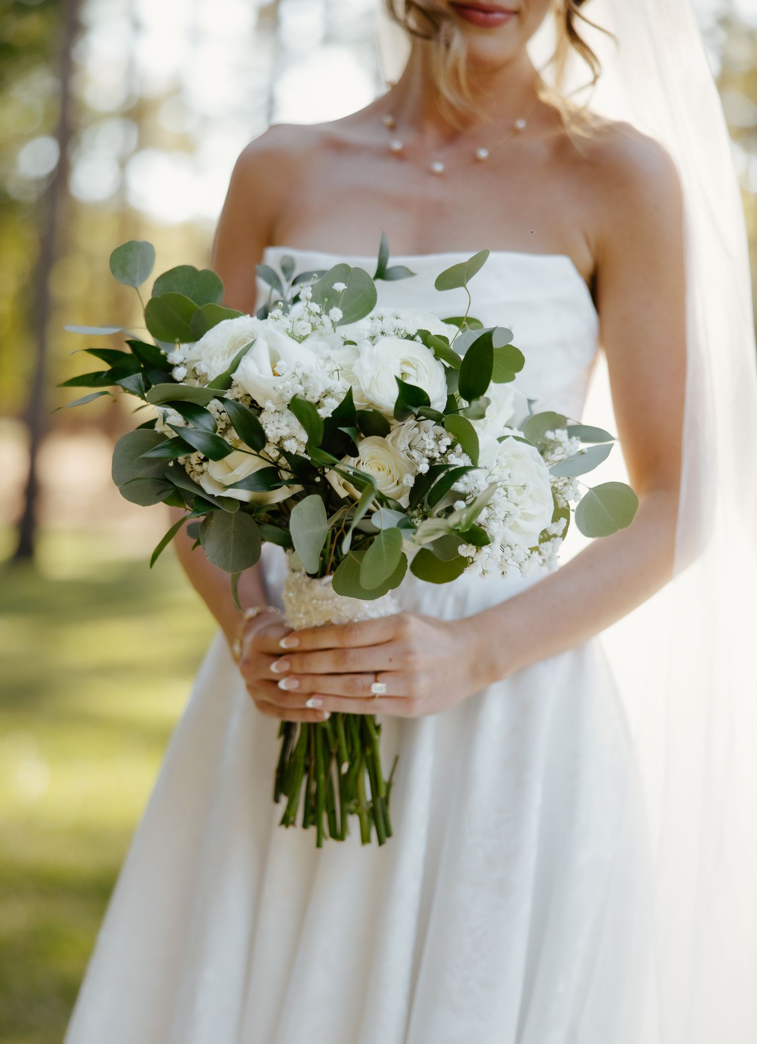Bride in strapless white gown holding elegant white rose and eucalyptus bouquet outdoors.