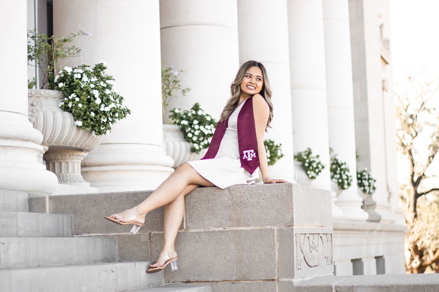 Young woman in maroon Texas A&M graduation sash sits on white stone steps near classical columns.