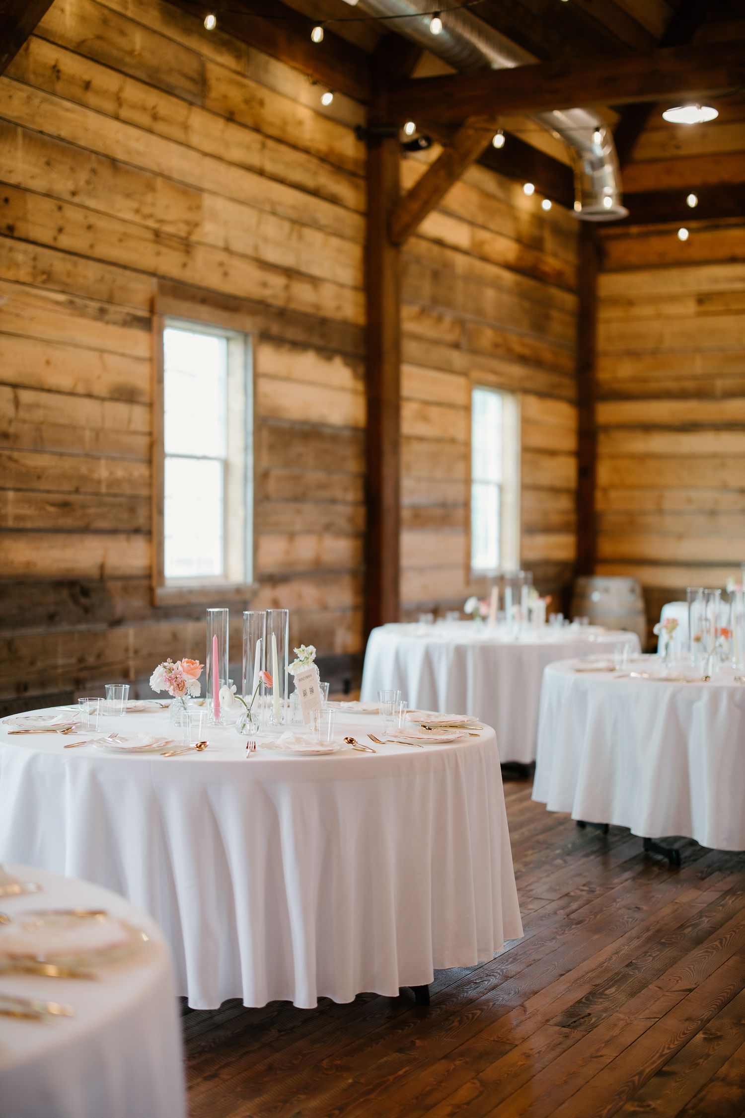 Elegant barn wedding reception with round tables draped in white linens and string lights overhead.