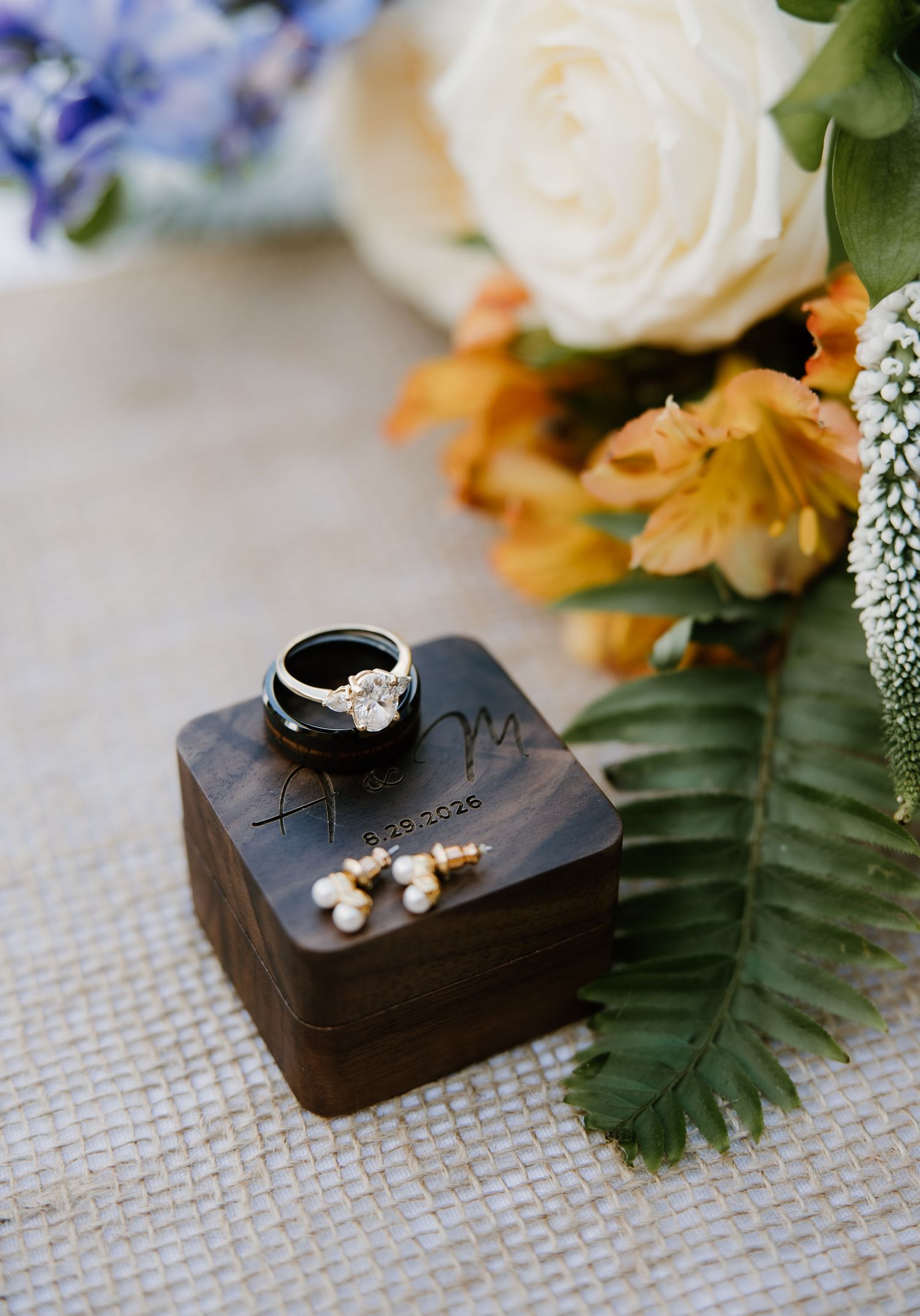 Wedding rings and pearl earrings displayed on engraved wooden ring box surrounded by white and orange florals.