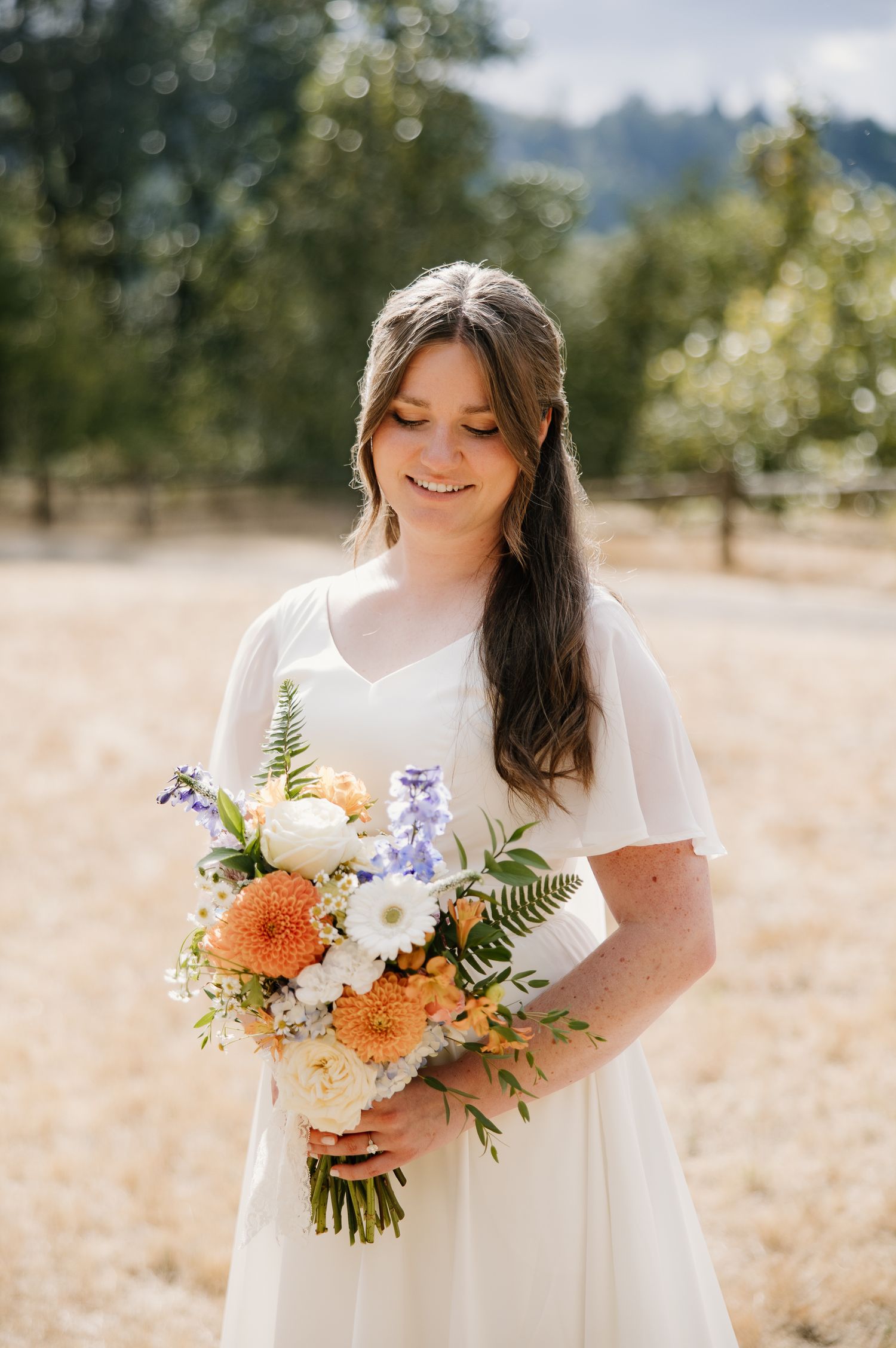 Smiling bride in white dress holding vibrant orange and white floral bouquet in sunny outdoor field.